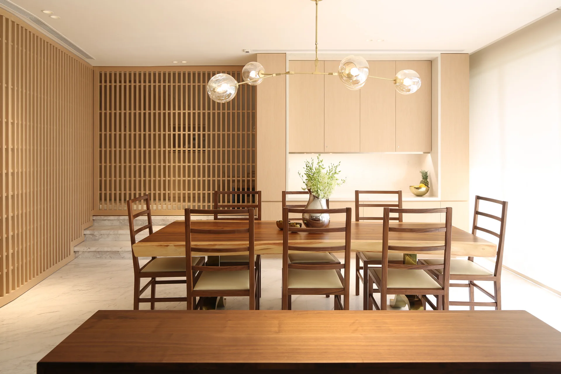 A modern dining room featuring a live-edge wooden table, matching chairs, a branching globe chandelier, and a vertical wooden slatted screen.