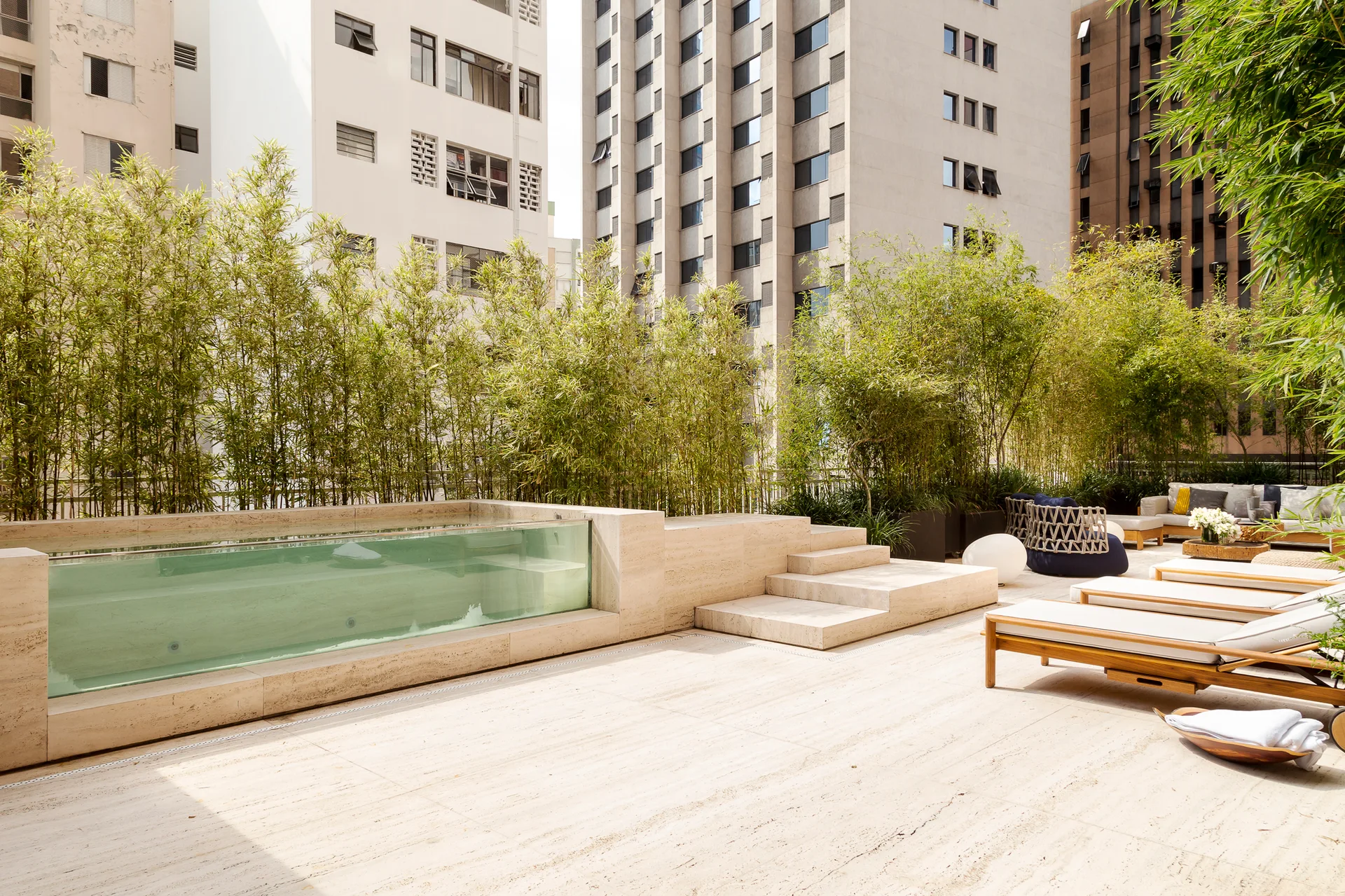 A raised travertine pool with a glass viewing wall sits on an urban rooftop deck lined with tall bamboo plants and wooden lounge chairs.