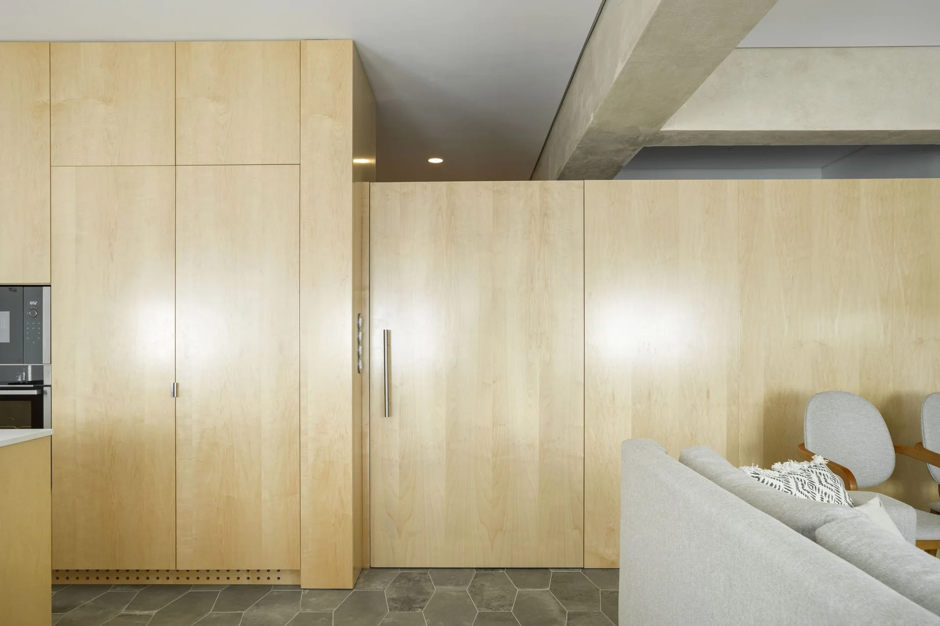 A minimalist interior featuring floor-to-ceiling light wood paneling with an integrated door, grey hexagonal floor tiles, and a concrete ceiling beam.