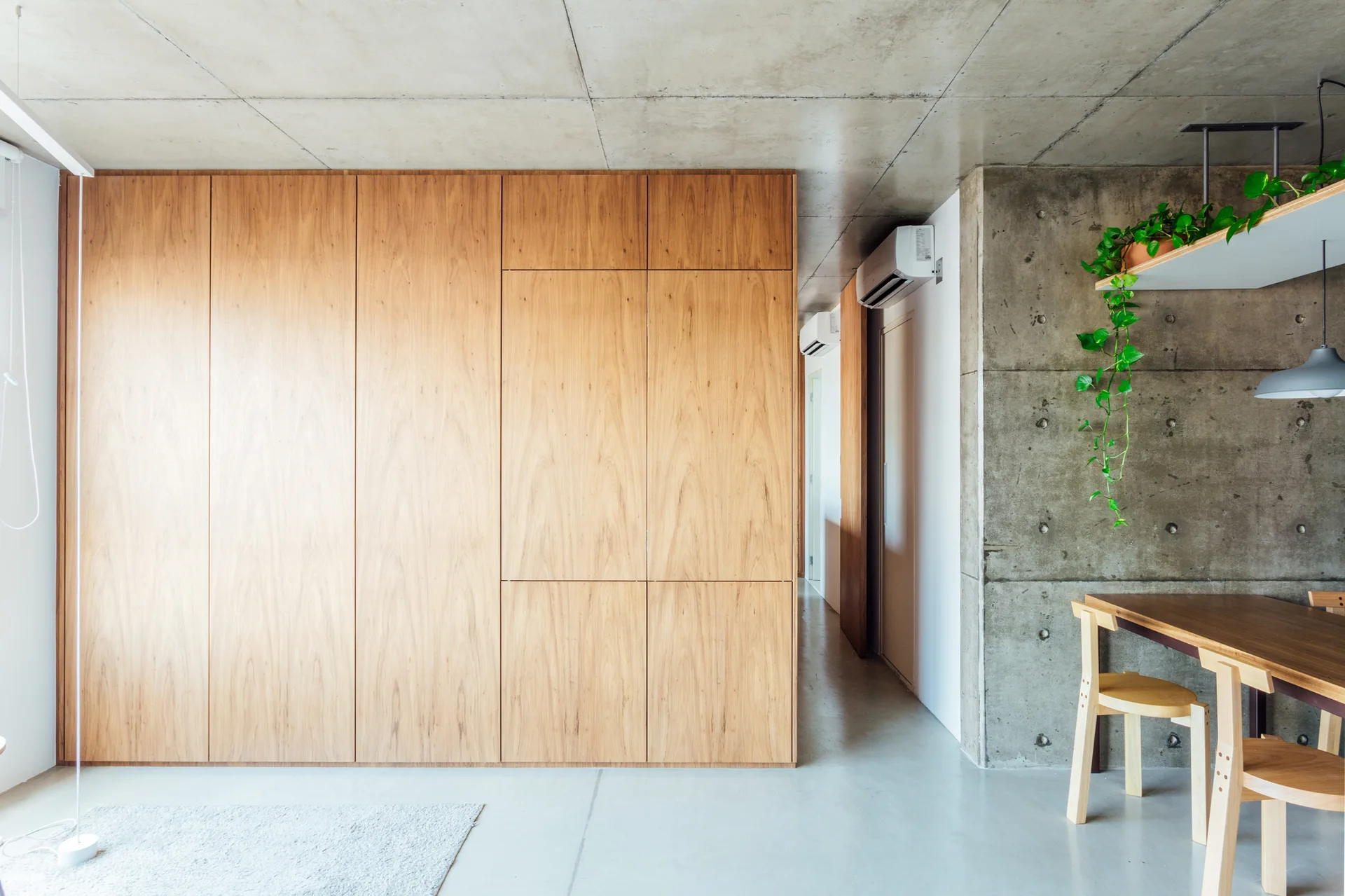 A minimalist dining room featuring a large wood-veneered storage wall, raw concrete ceiling and walls, and a simple dining table set.