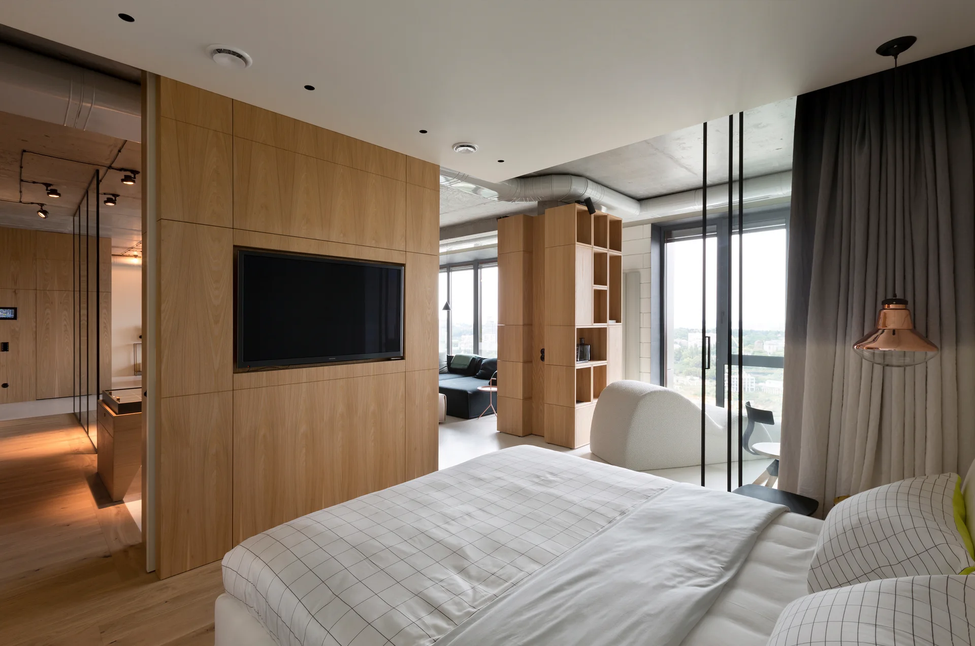 A modern bedroom featuring a bed with a grid-patterned duvet, a wood-paneled wall with a mounted television, and an exposed concrete ceiling.