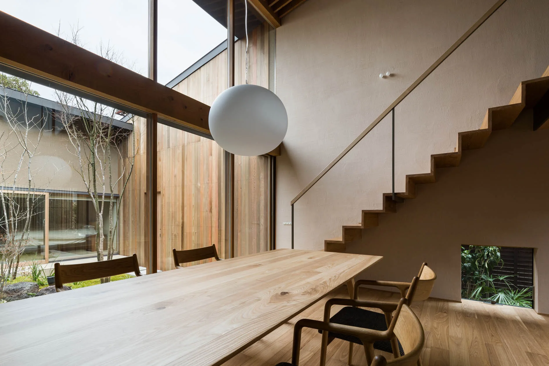 A minimalist dining room features a long wooden table, a large white globe pendant, and a floating wooden staircase next to tall glass walls.