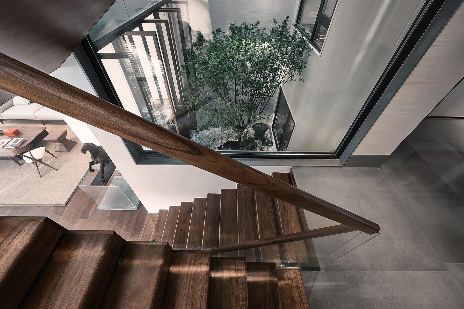 A high-angle view of a dark wood staircase with glass railings overlooking a glass-walled internal atrium featuring a live green tree.