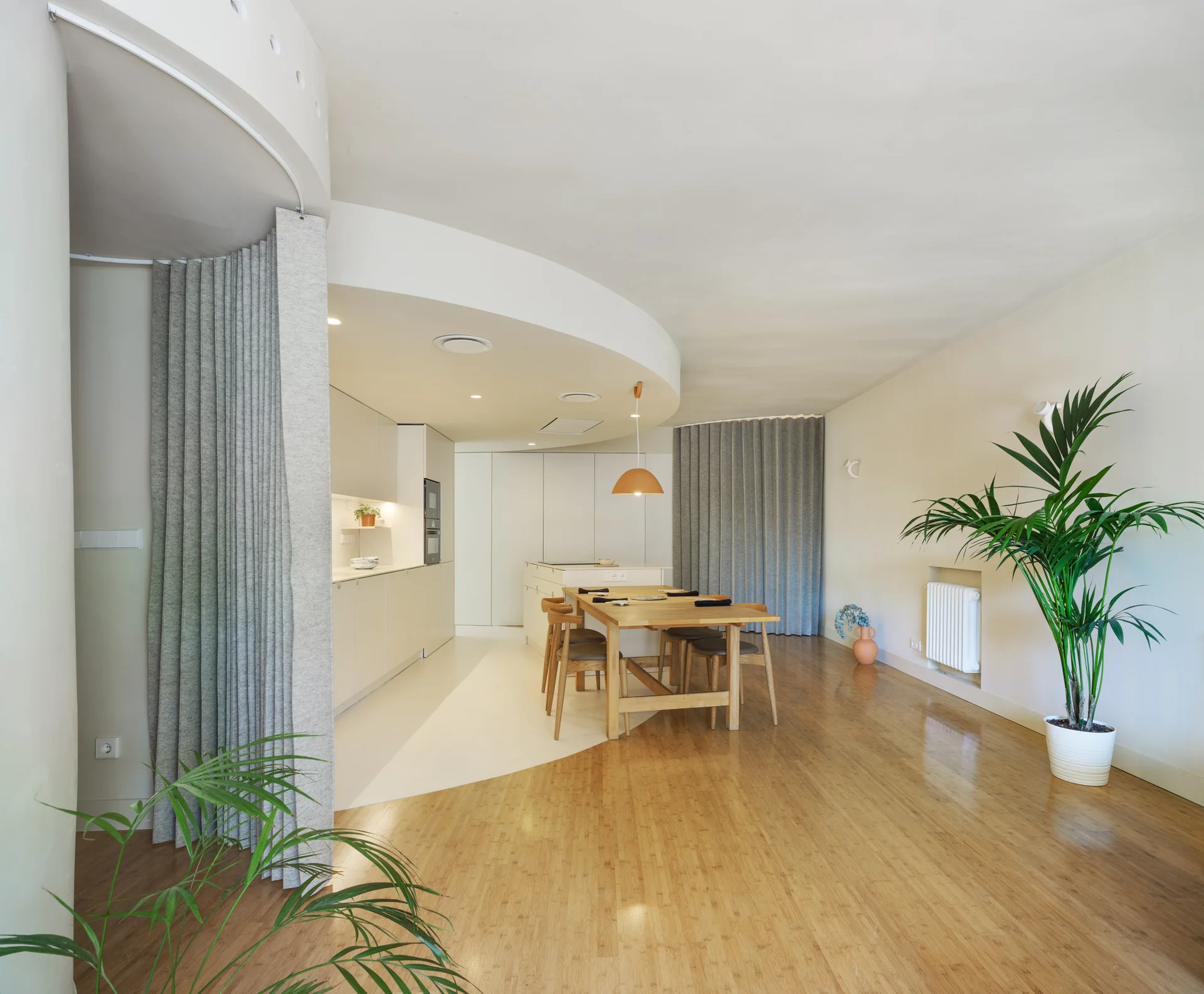 A modern dining area featuring a wooden table, light bamboo flooring, and grey pleated curtain room dividers near a kitchen with curved ceiling architecture.