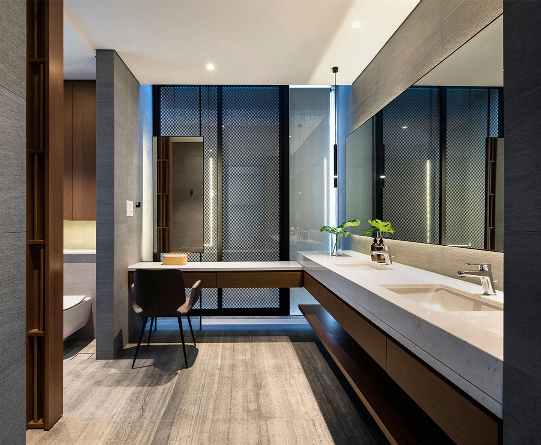 A modern bathroom featuring a long white double vanity with wooden cabinets, a matching makeup desk with a dark chair, and textured gray walls.
