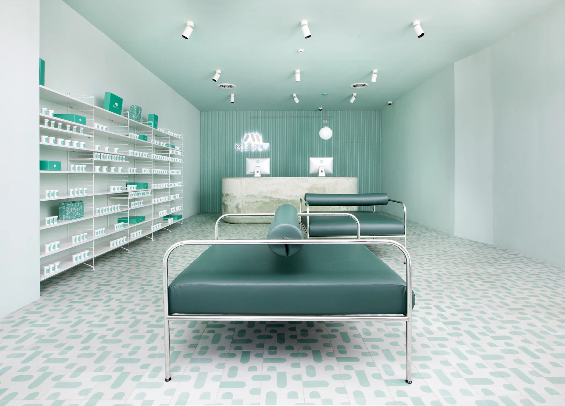 A minimalist mint green pharmacy interior featuring geometric patterned tile floors, white open shelving, a stone-like desk, and dark green leather benches.