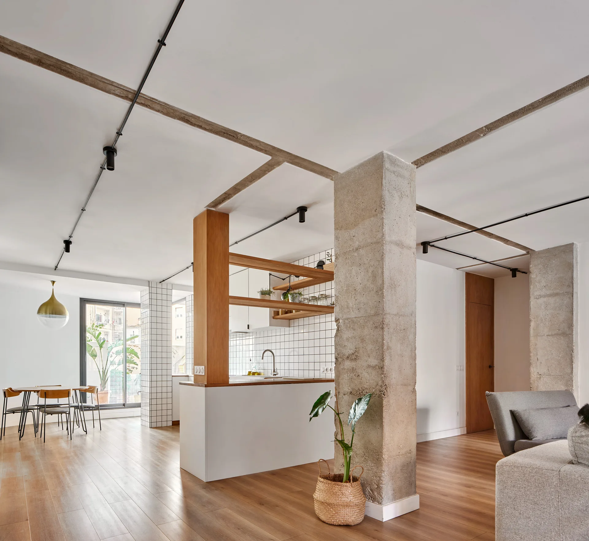An open-plan residential interior features raw concrete structural columns, wood flooring, a white kitchen island, and square grid tiled walls.