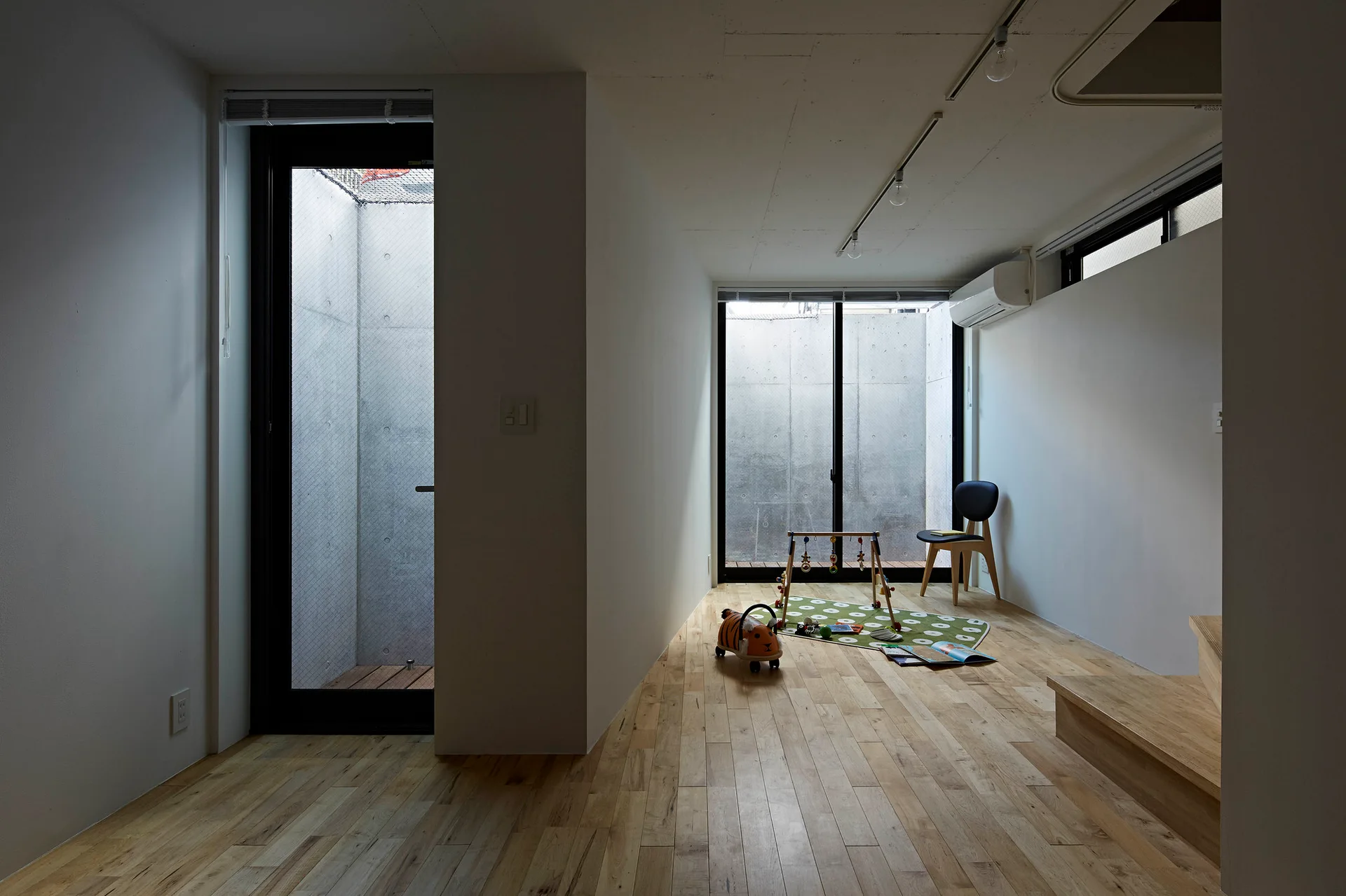 A minimalist children's playroom features light wood plank floors, white walls, and black-framed glass doors that open to concrete-walled lightwells.
