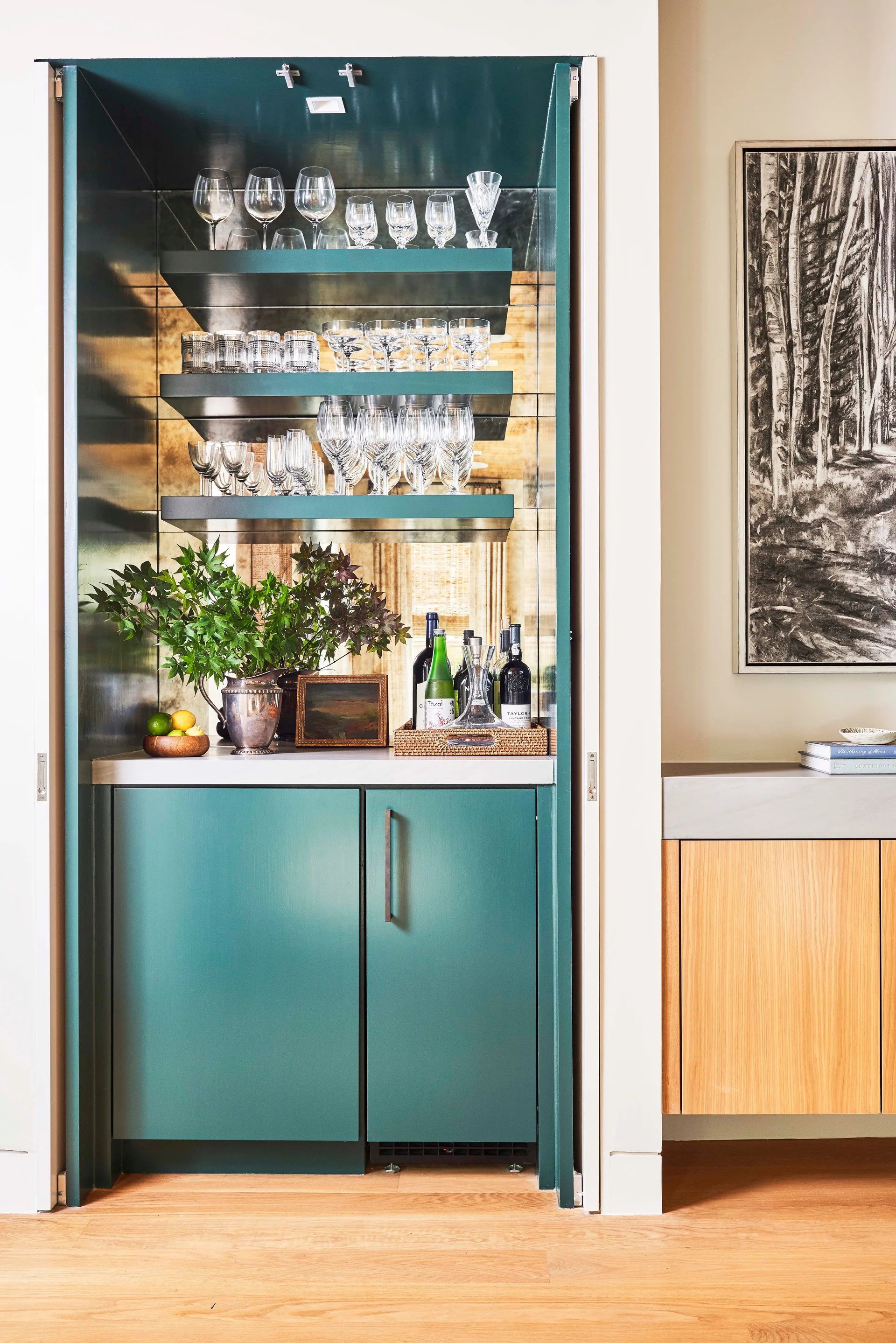 A built-in teal home bar features open floating shelves with glassware, a mirrored backsplash, and lower cabinets on light wood floors.