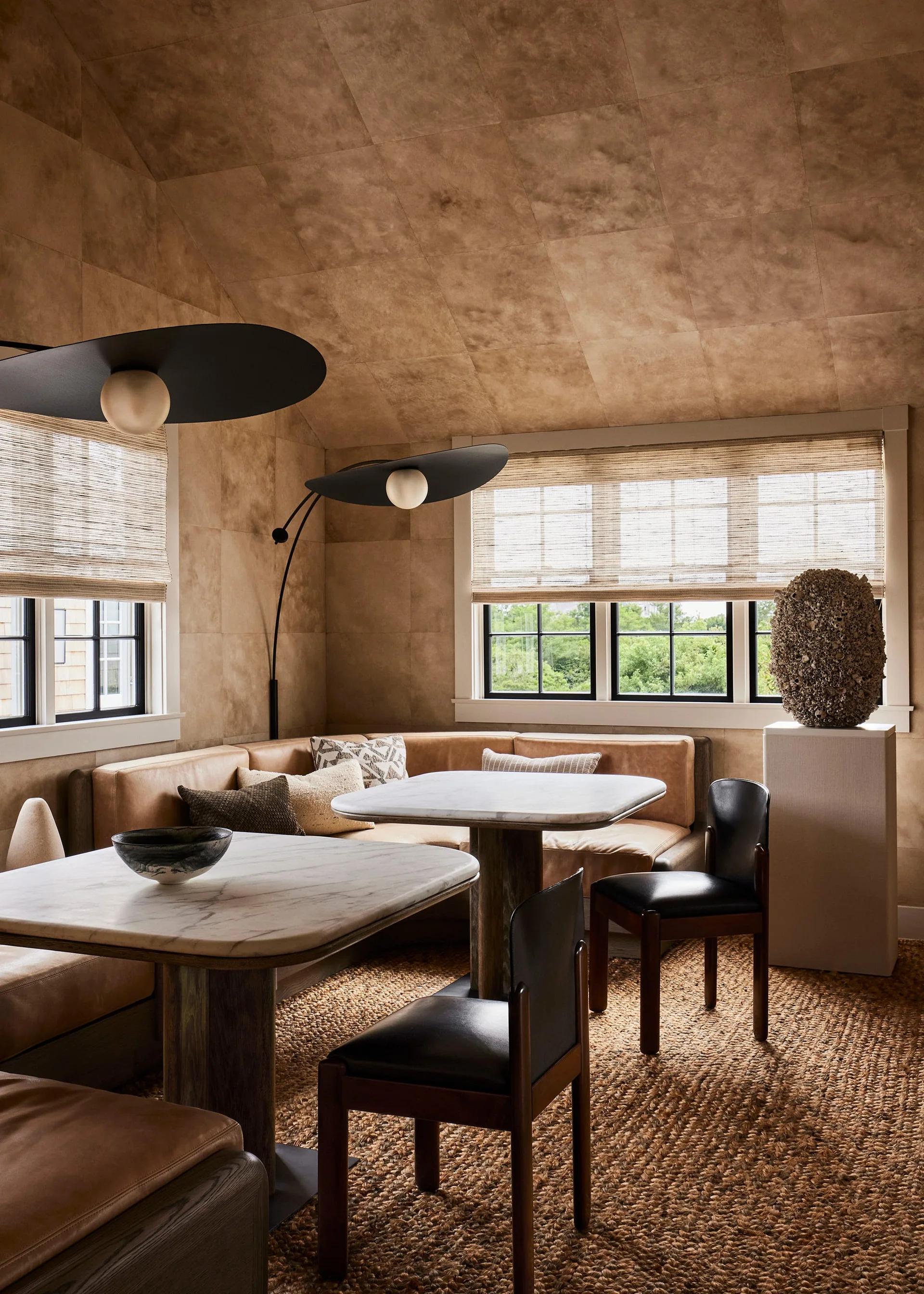 A dining nook features a tan leather corner booth, two marble tables, and black chairs beneath a sloped ceiling lined with square textured wall tiles.