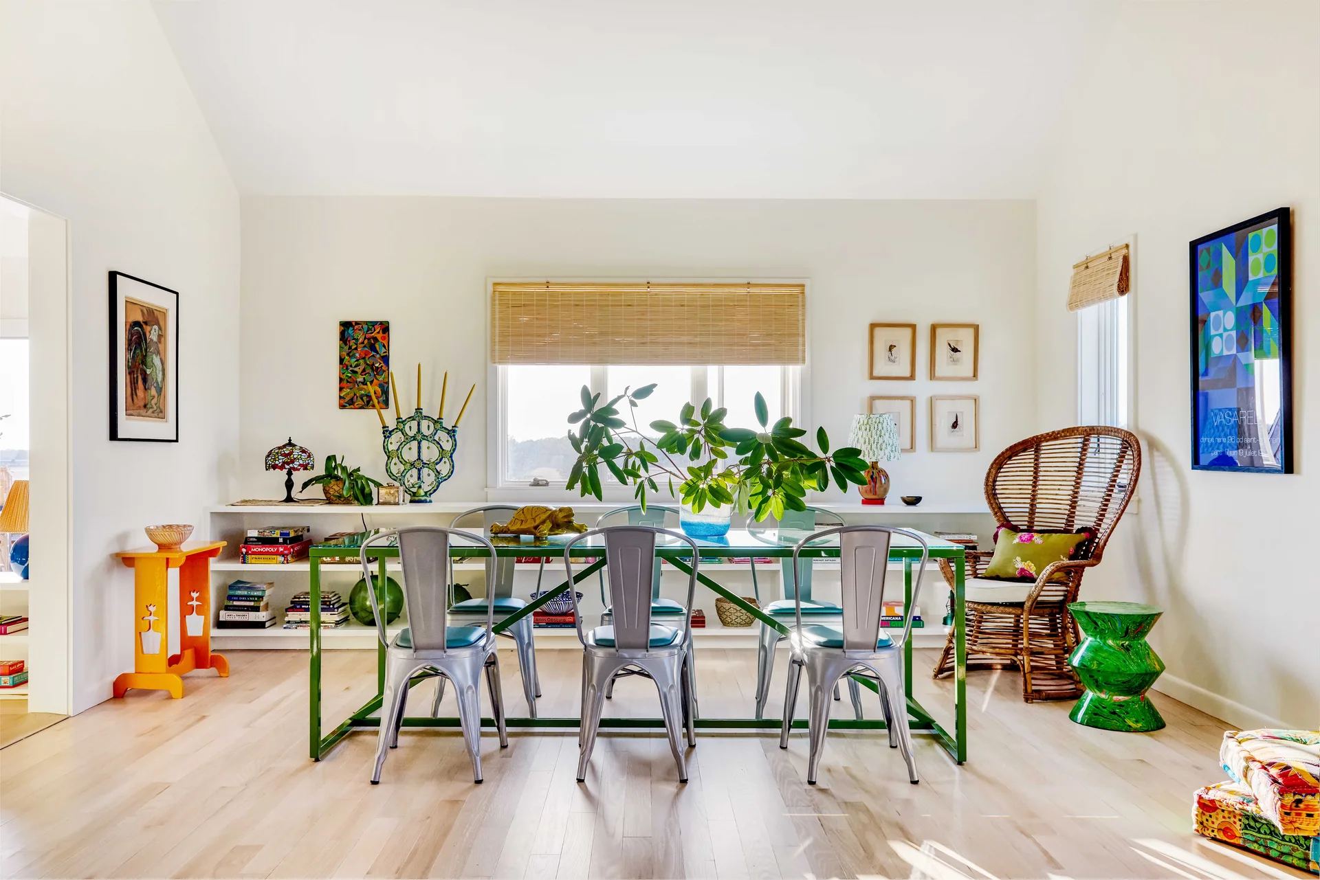 A bright dining room featuring a green metal dining table, silver bistro chairs, a rattan peacock chair, and low white built-in bookshelves.