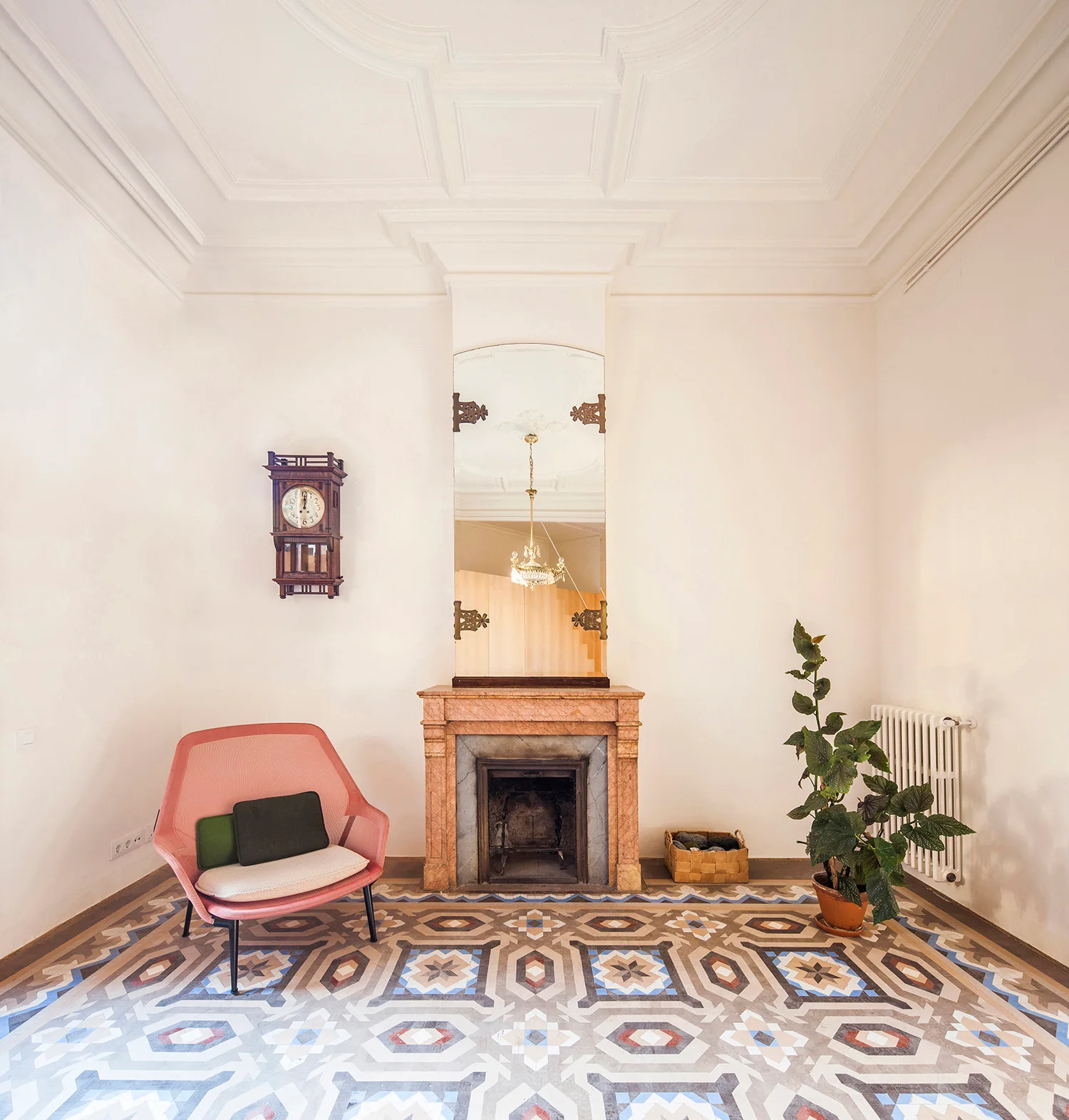 A bright room features colorful geometric floor tiles, a pink armchair, an antique wall clock, and a stone fireplace with a tall mirror above it.