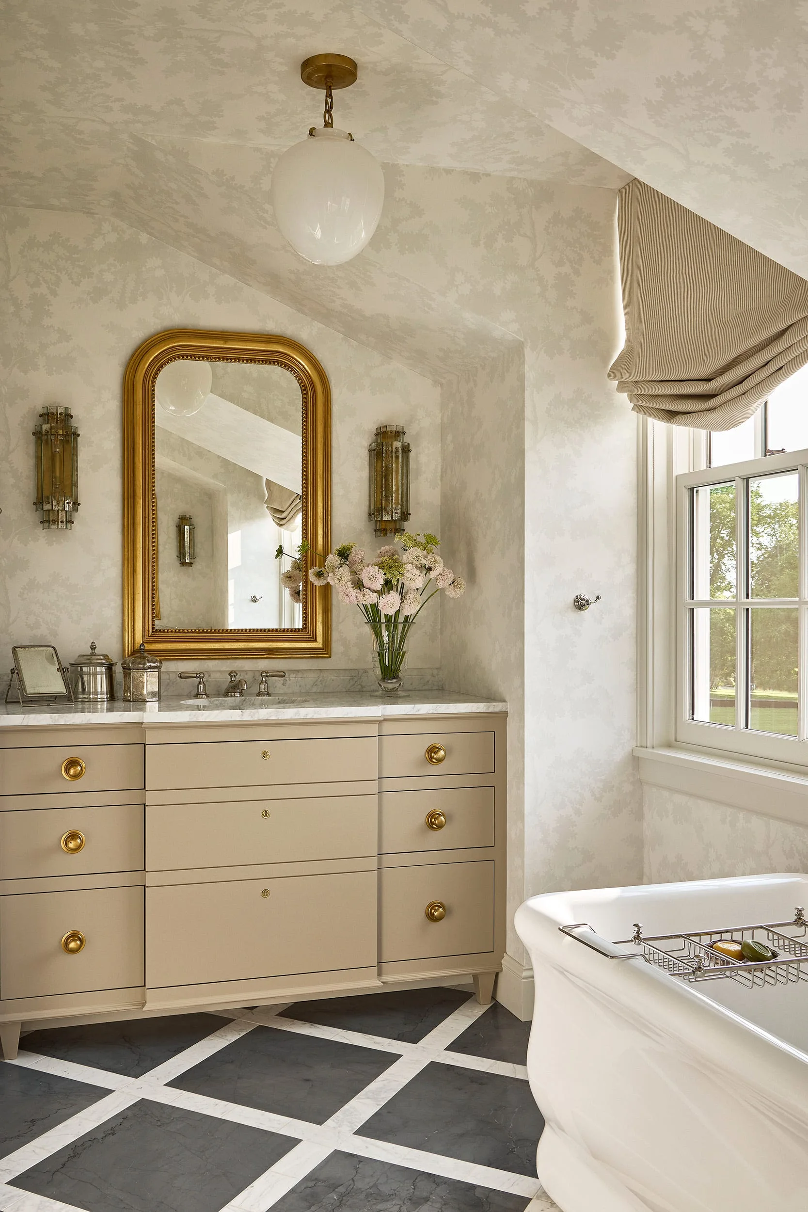 A traditional bathroom features a taupe vanity with a marble countertop, an arched gold-framed mirror, and checkered stone flooring next to a tub.