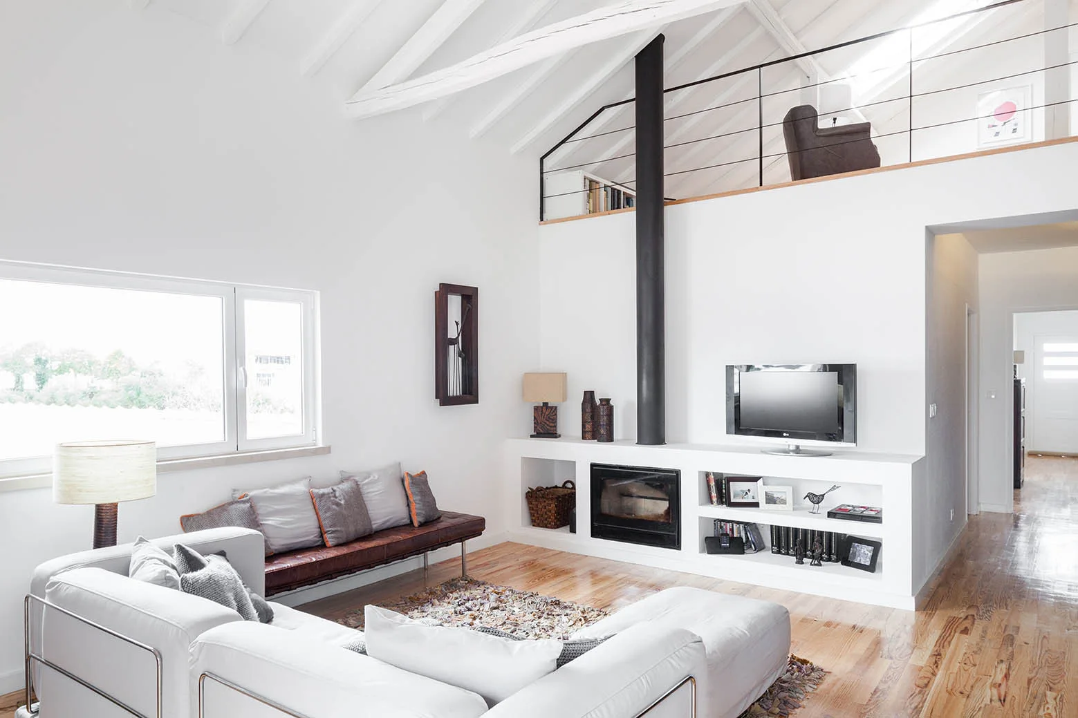 A double-height white living room featuring a vaulted ceiling with exposed beams, a built-in fireplace with a tall black flue, and a mezzanine loft.