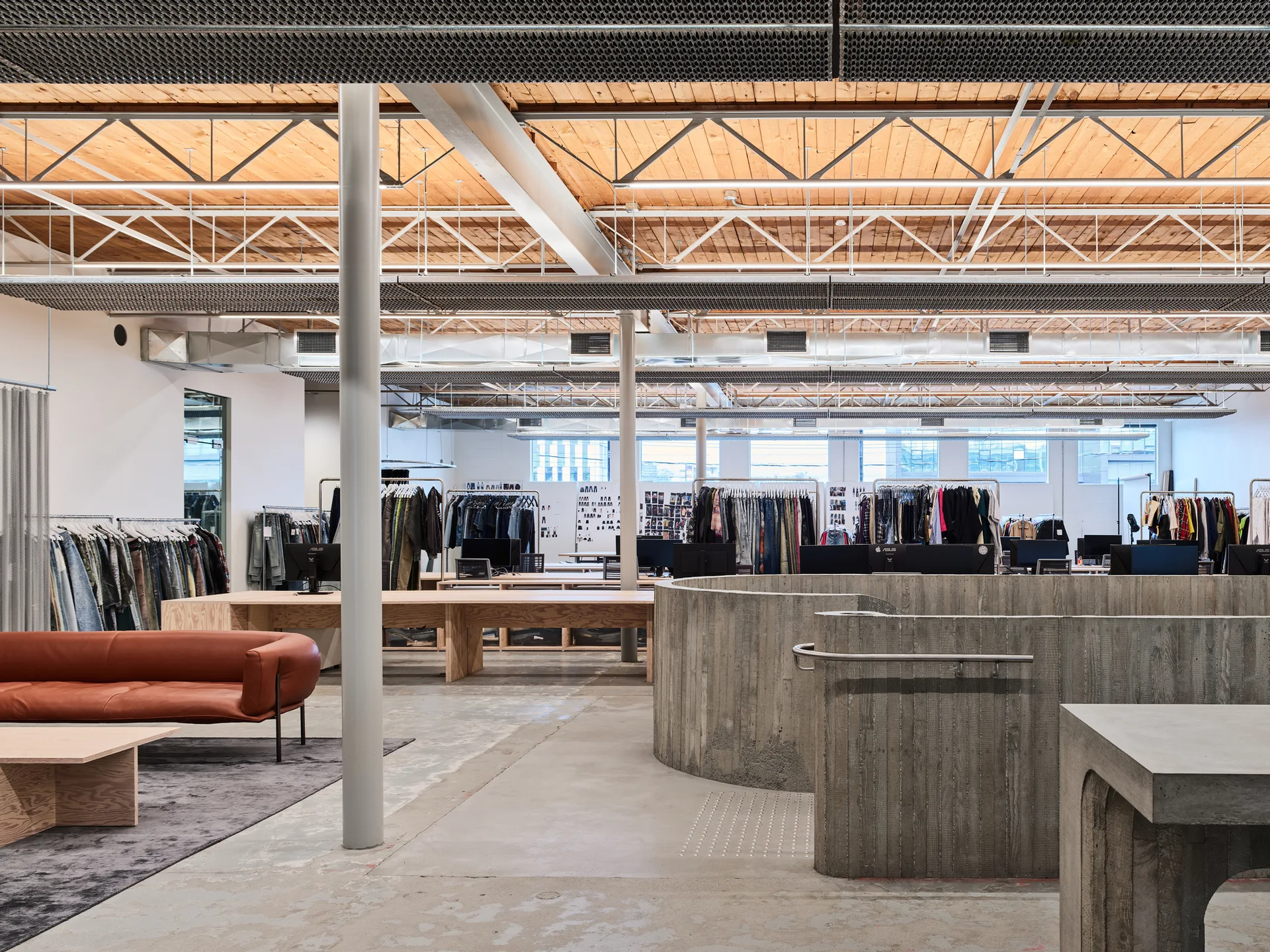 An open industrial workspace featuring a curved concrete wall, exposed timber ceilings, long wood desks, and clothing racks.