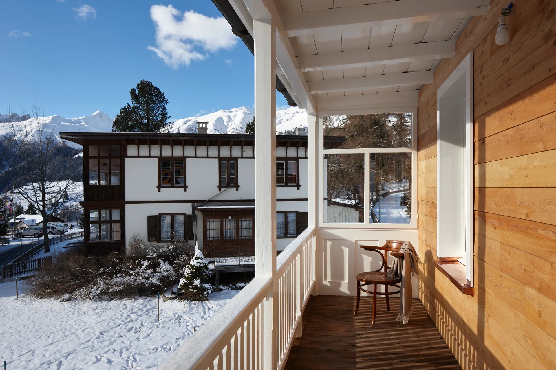 A covered wooden balcony with light pine siding features a single bentwood chair with a draped blanket overlooking a snowy alpine landscape.