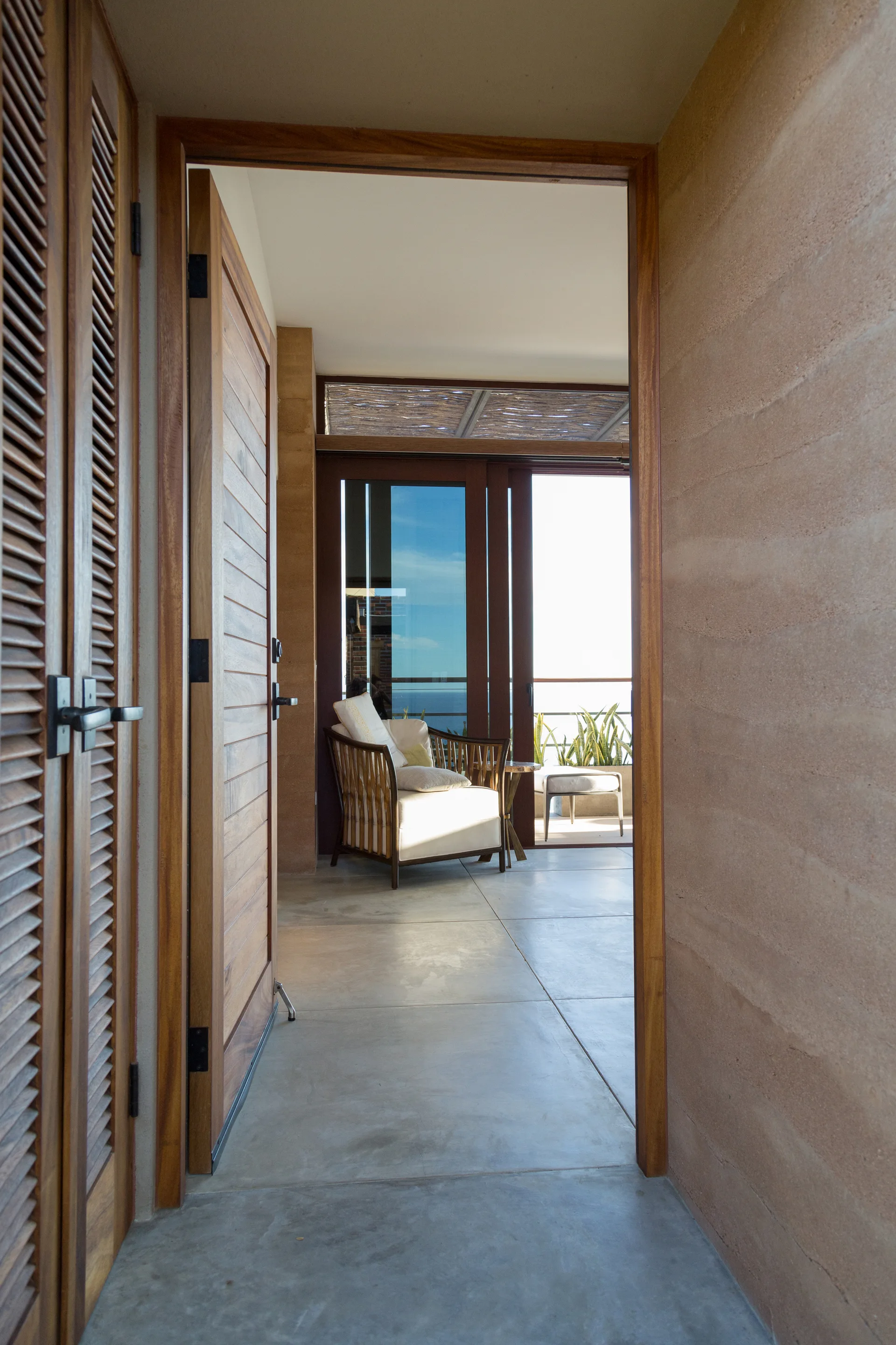 View down a concrete corridor with a textured rammed earth wall and louvered wooden doors leading to a bright living area with an armchair.