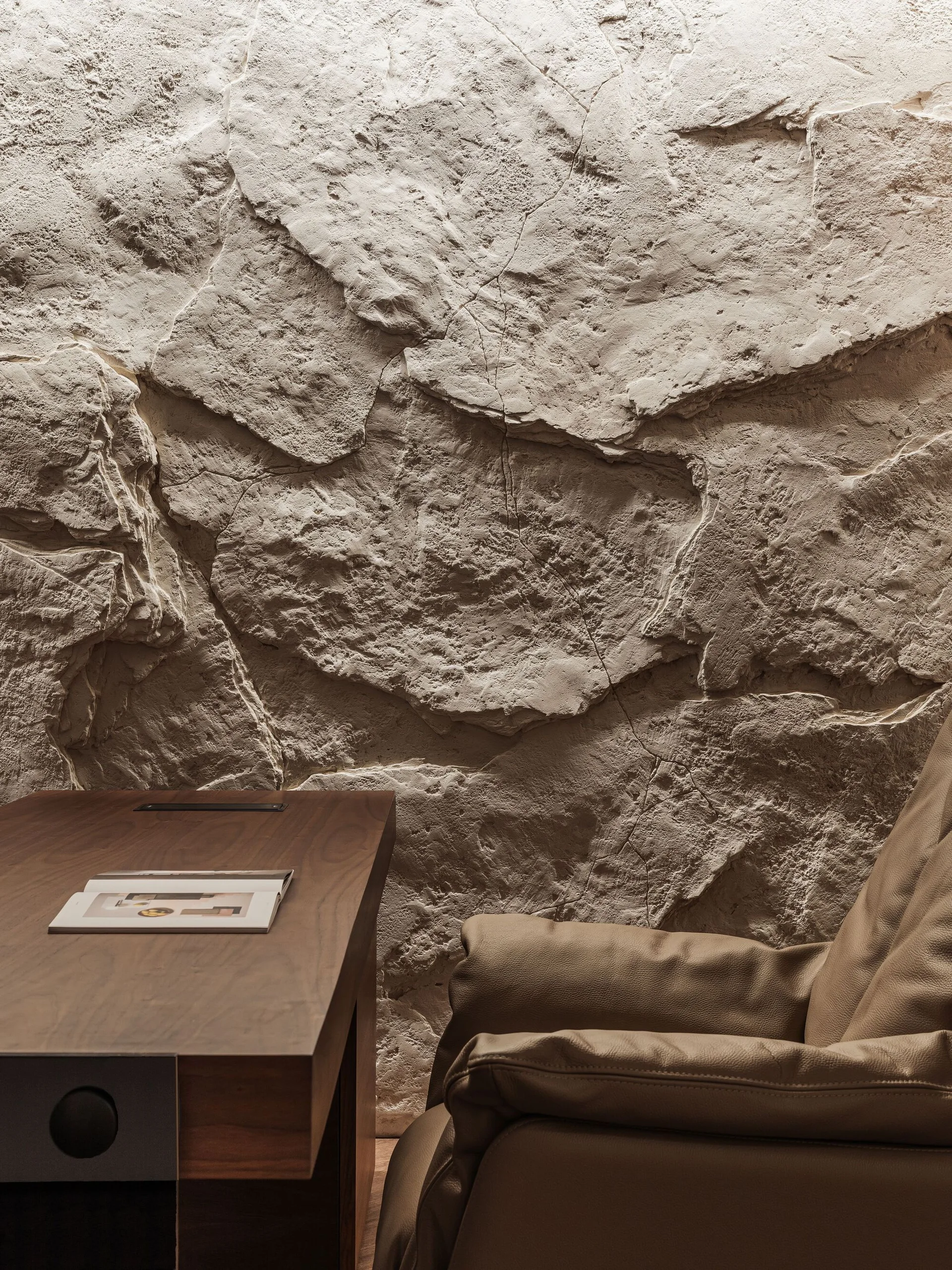 A rough beige stone accent wall stands behind a dark wood desk with an open book and a tan leather armchair.