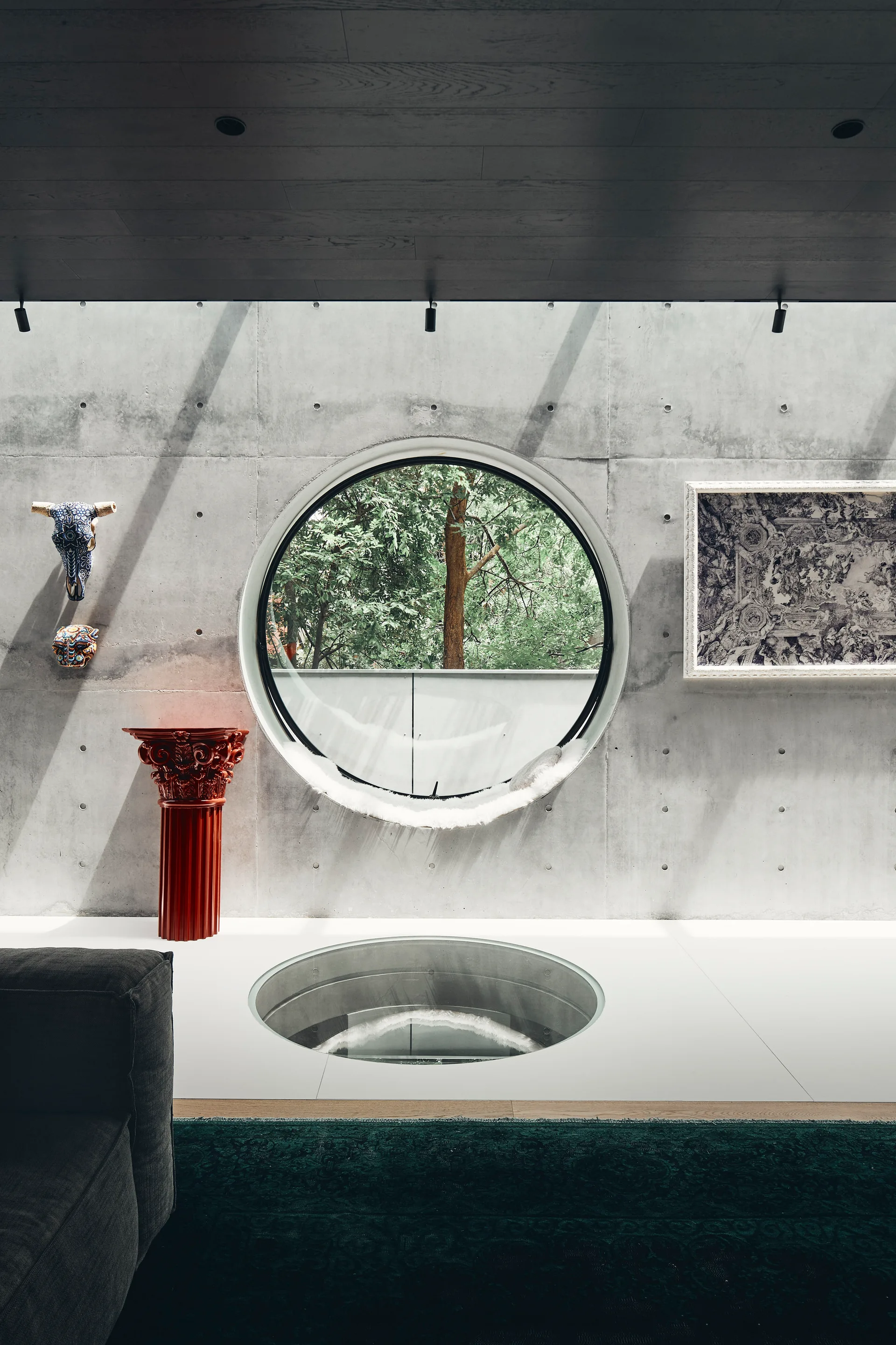A modern interior features an exposed concrete wall with a large circular window, a circular floor window, and a bright red classical pedestal.