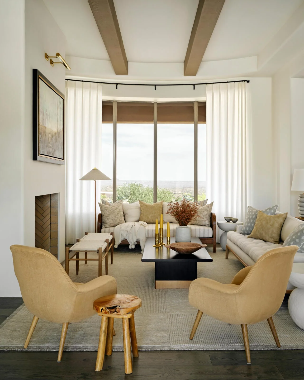 A symmetrical living room features tan armchairs, neutral sofas, a dark wood coffee table, and large windows flanked by white curtains under ceiling beams.