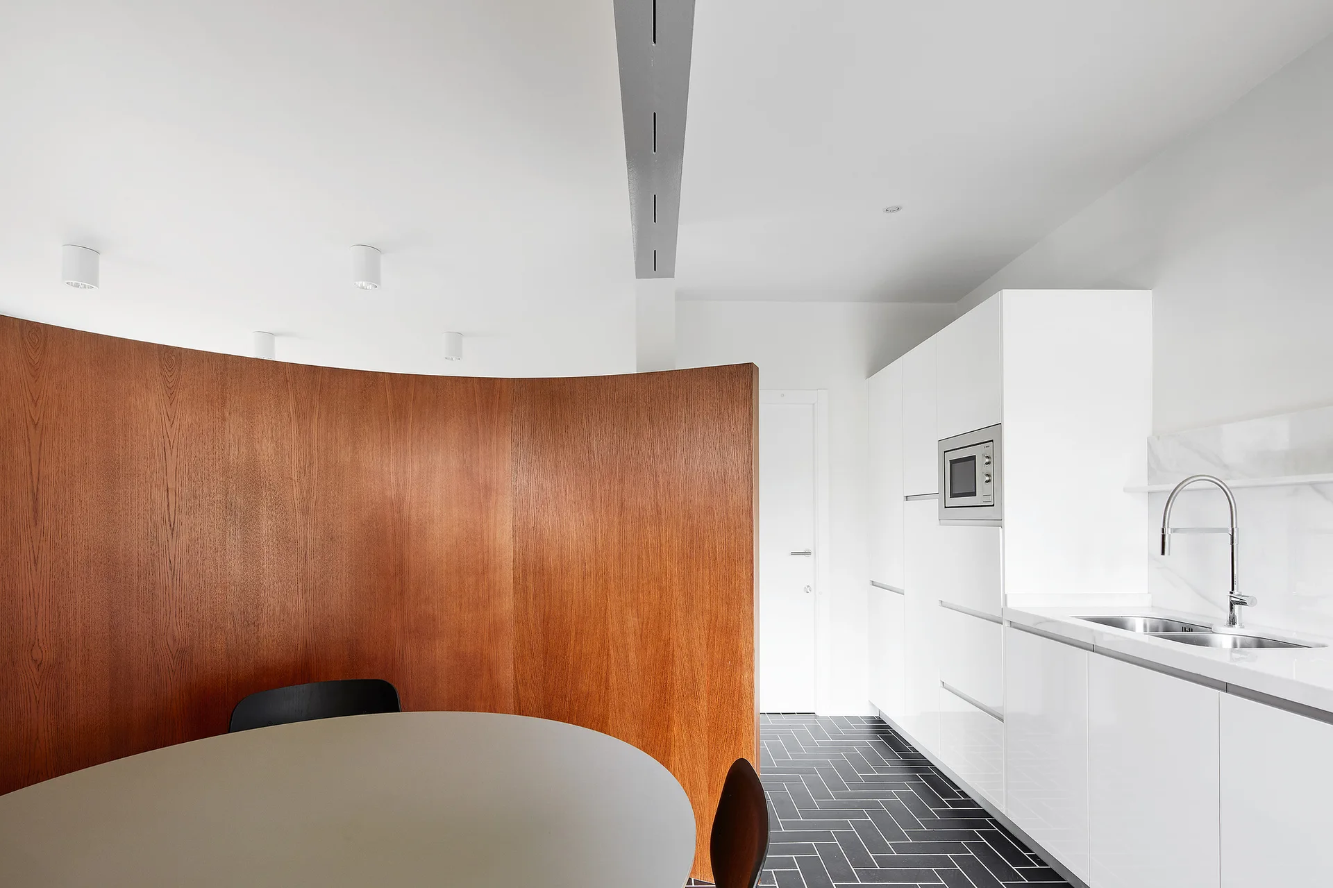 A modern white kitchen with handleless cabinets, a curved wooden partition wall, black herringbone floor tiles, and a round dining table.