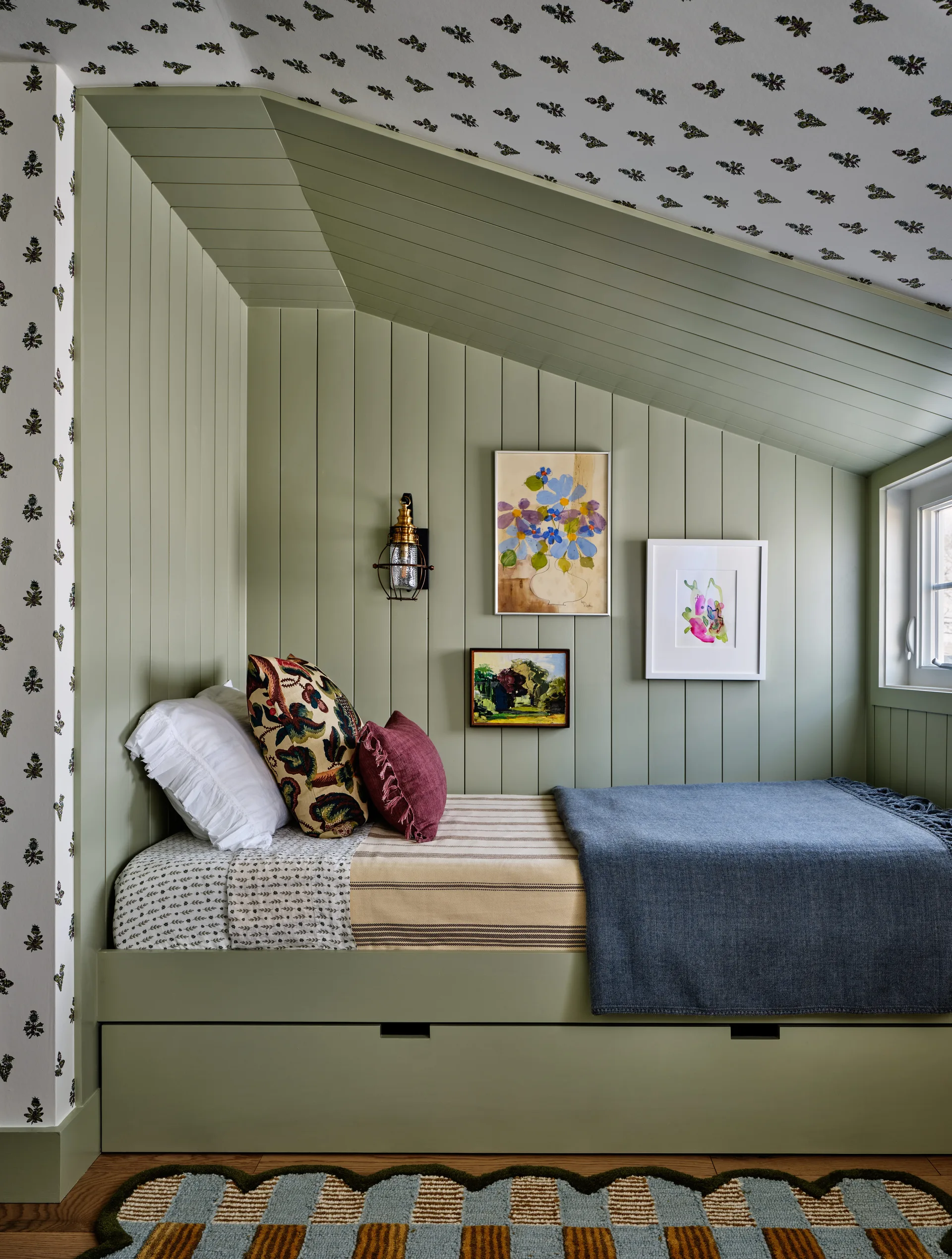 A built-in sage green daybed sits under a sloped ceiling in a cozy bedroom alcove featuring vertical wood paneling and floral wallpaper.