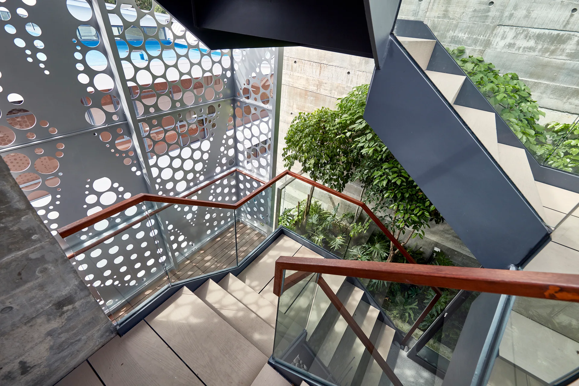 A modern interior staircase features light wood treads, clear glass railings, wood handrails, and a metal screen with circular perforations.