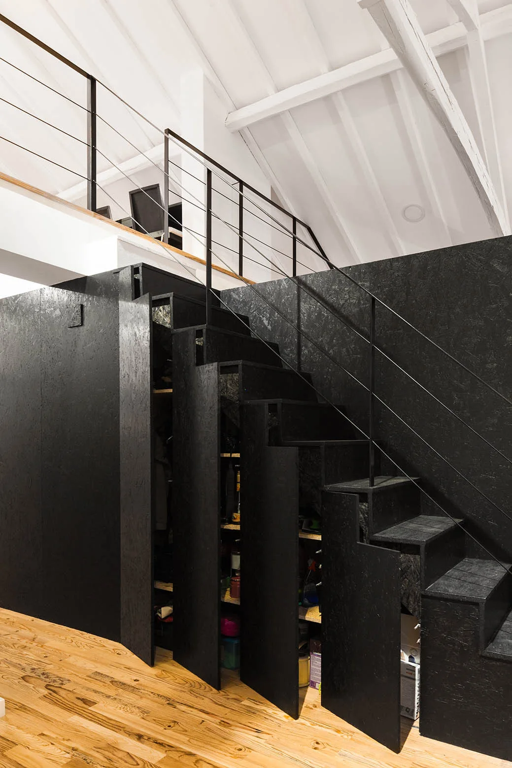 A modern black staircase with integrated under-stair storage drawers stands on light wood floors beneath a white vaulted ceiling.