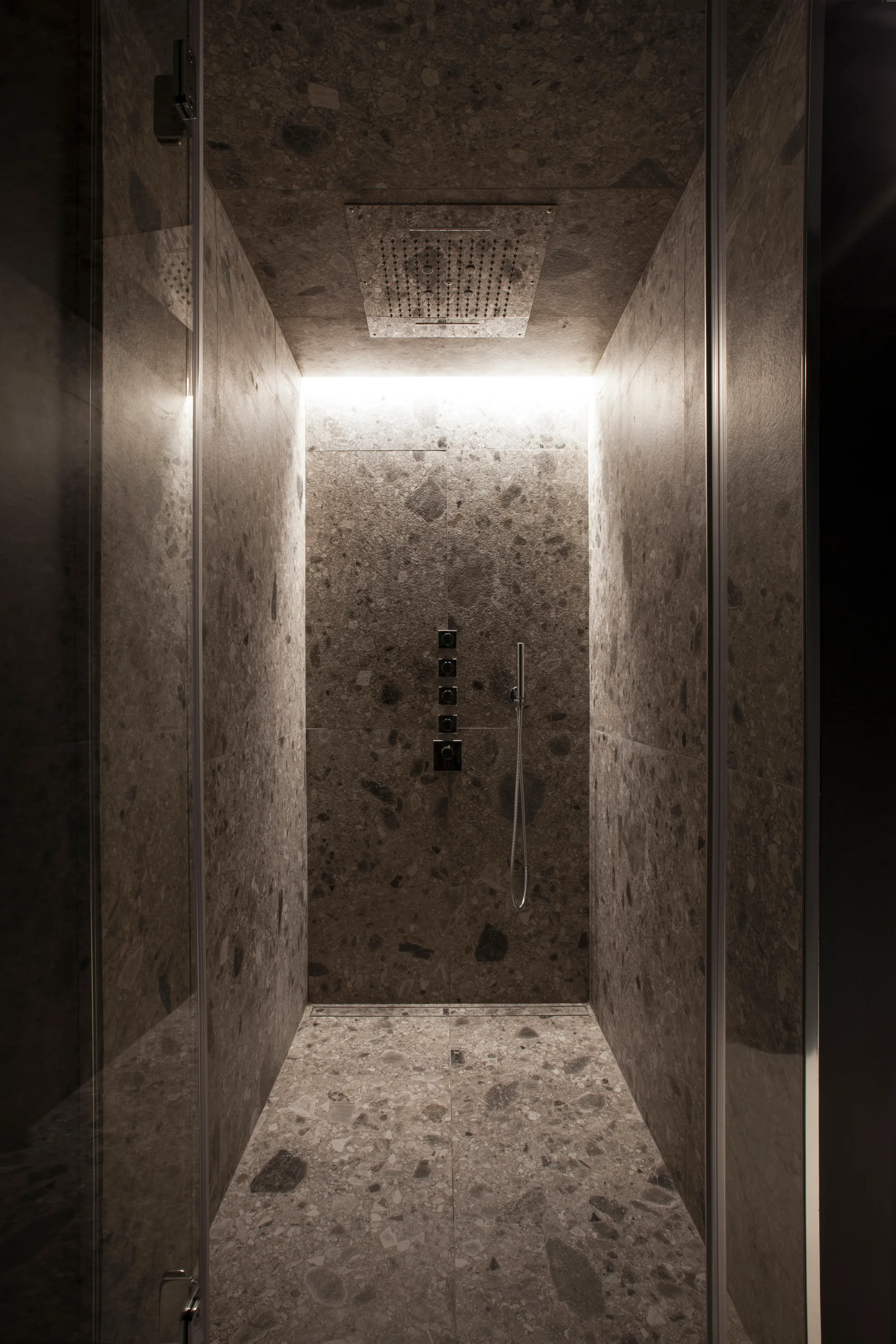 A modern walk-in shower with brown speckled stone tiles covering the walls, floor, and ceiling, featuring integrated cove lighting and a rain shower head.