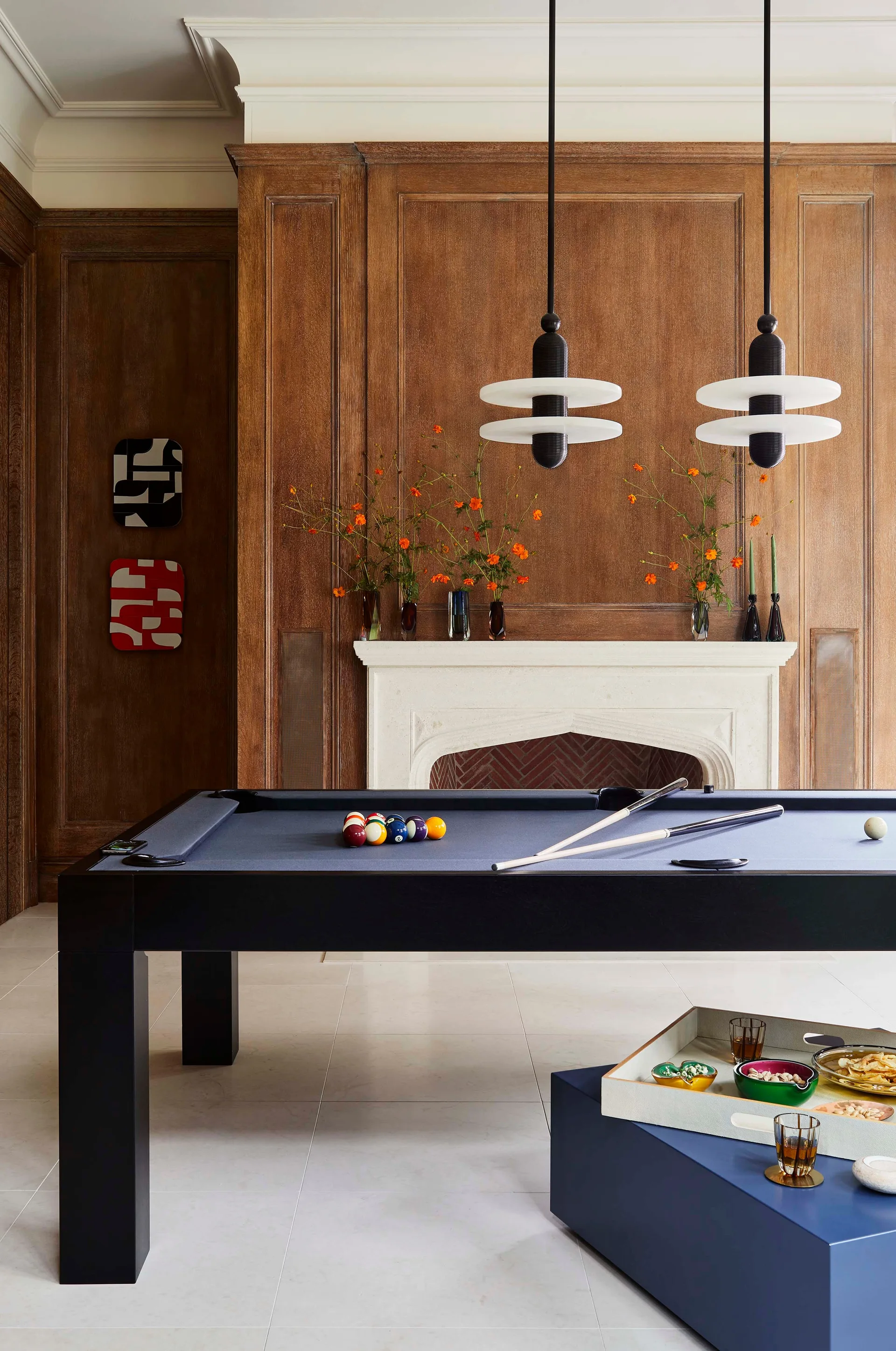 A navy pool table stands in a game room with rich wood wall paneling, a stone fireplace, and two modern pendant lights overhead.