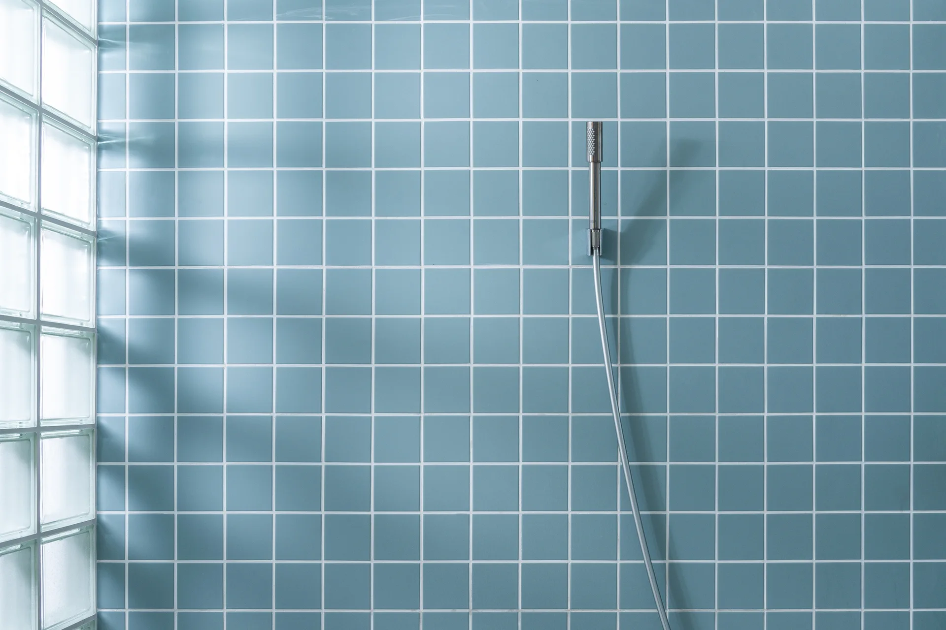 A light blue square-tiled shower wall features a sleek silver handheld wand next to a wall of translucent glass blocks.