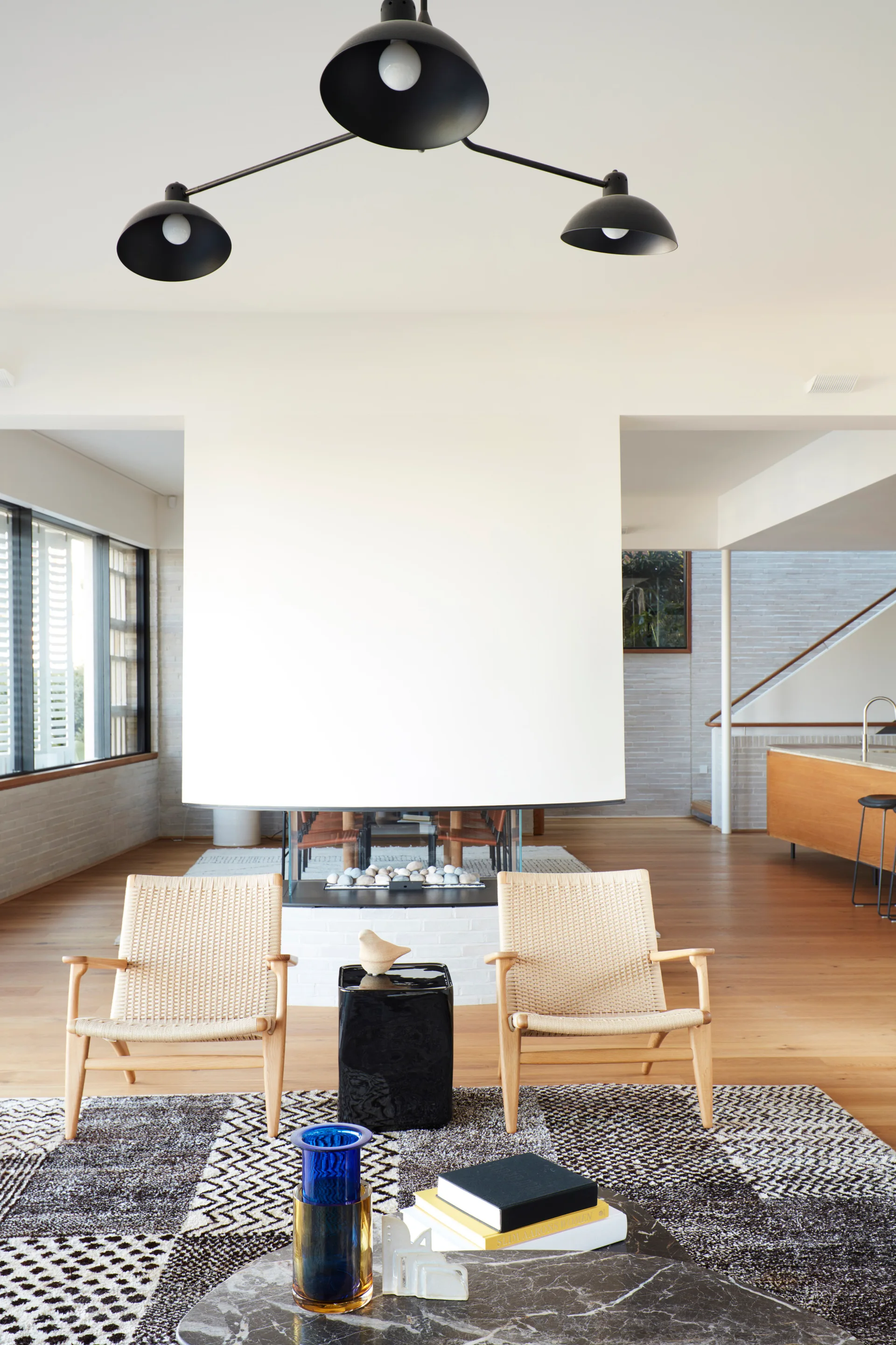 Two woven wood armchairs sit on a patterned rug facing a central white double-sided fireplace in a modern, light-filled living room.