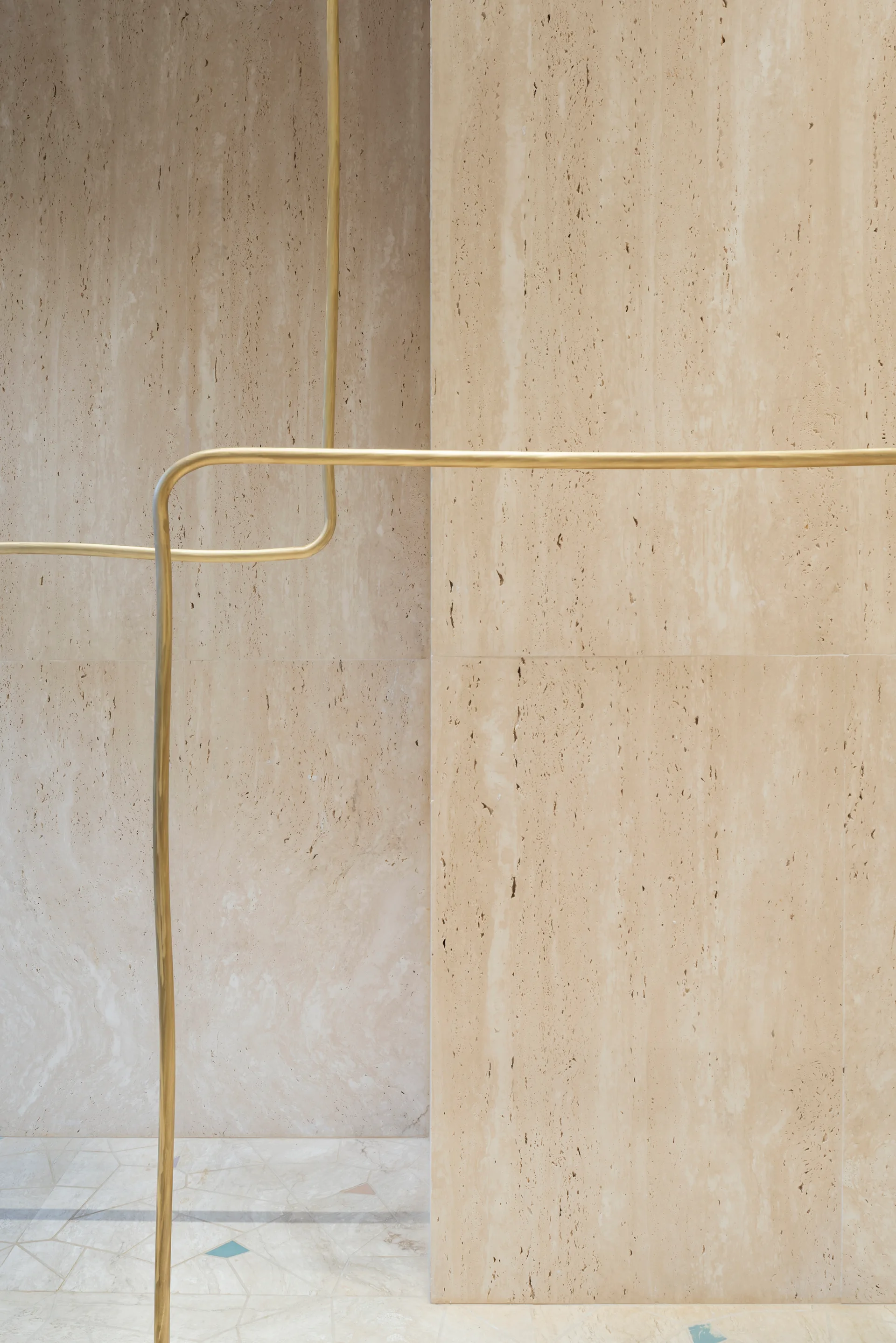 Honed beige travertine stone wall panels are paired with a slim curved brass railing and a patterned stone tiled floor.