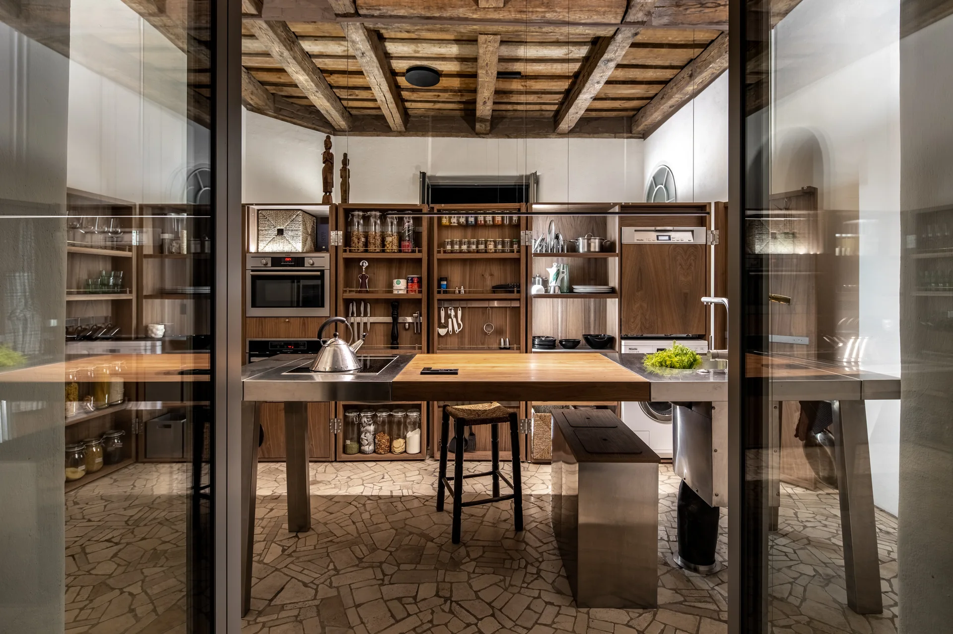 A kitchen with a rustic wooden beamed ceiling, tall wood cabinetry, an island with a stainless steel frame and wood top, and flagstone floor.