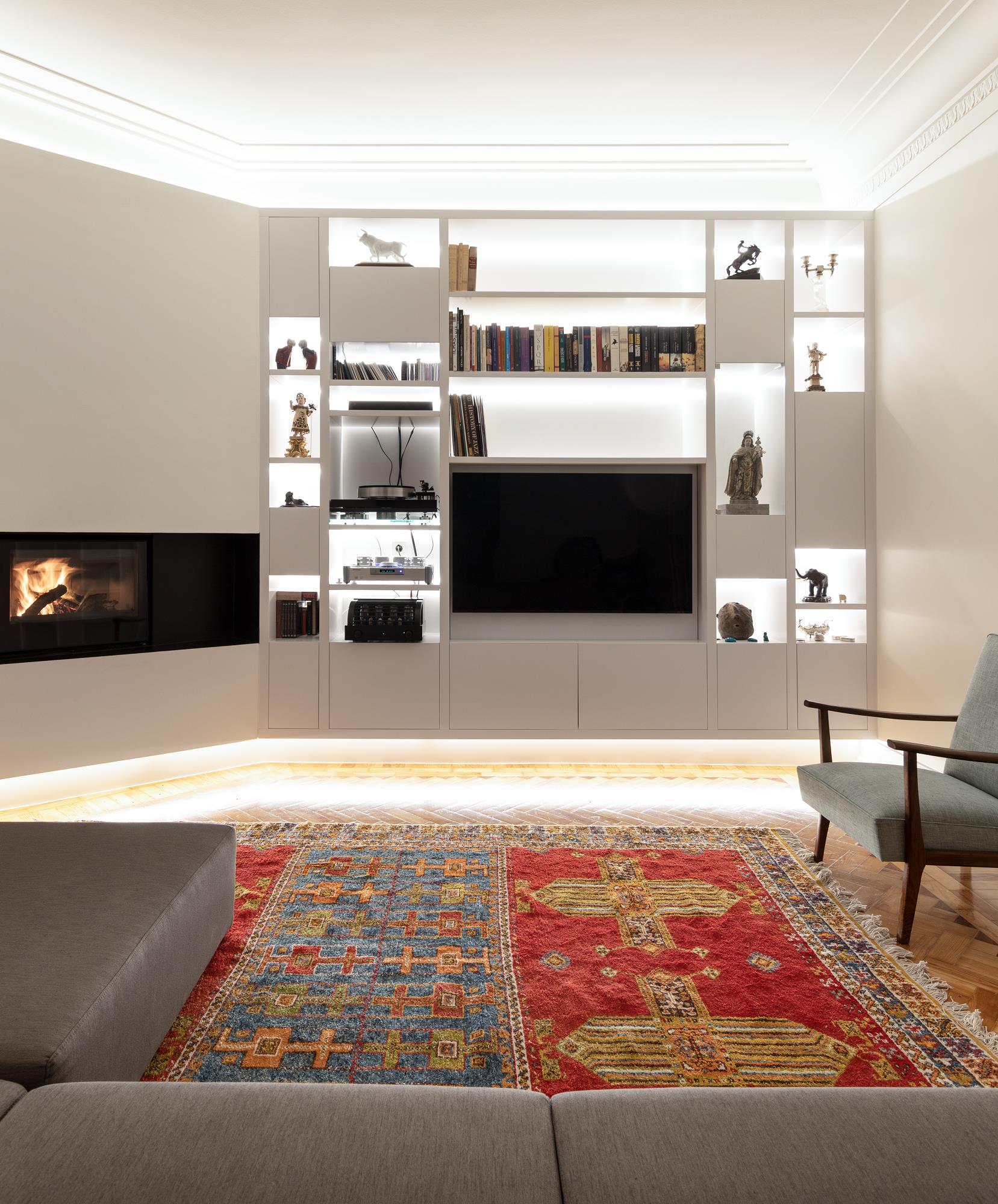 A modern living room features a white custom shelving unit with integrated lighting, a black television, a corner fireplace, and a vibrant red area rug.
