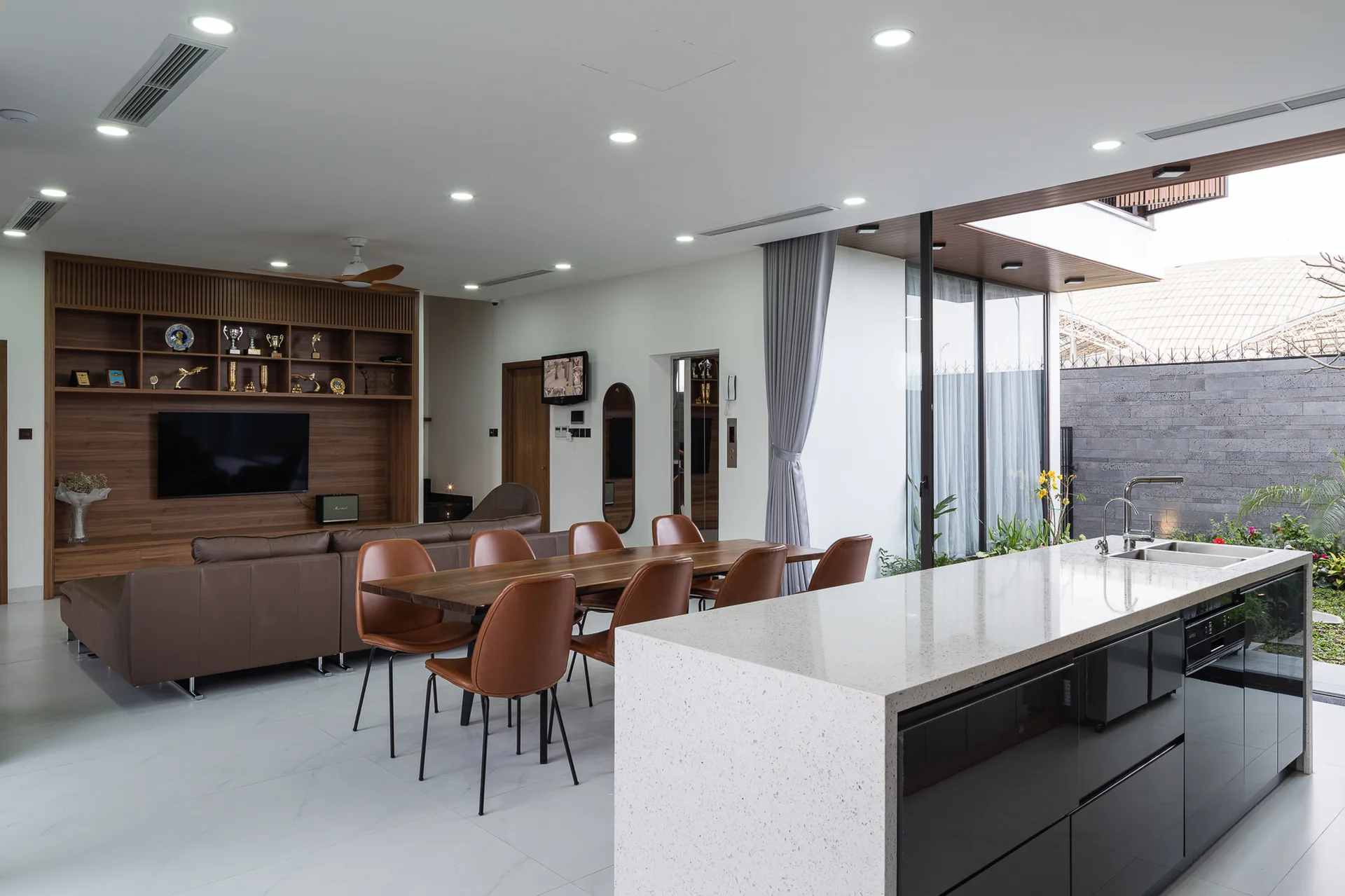 An open-plan interior showing a white stone kitchen island, a wood dining table with brown leather chairs, and a wood-paneled wall unit in the background.