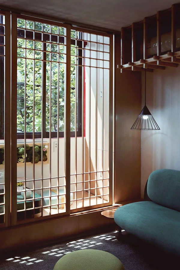 A teal curved sofa sits near a large wooden lattice window screen with natural light filtering through next to a hanging pendant lamp.