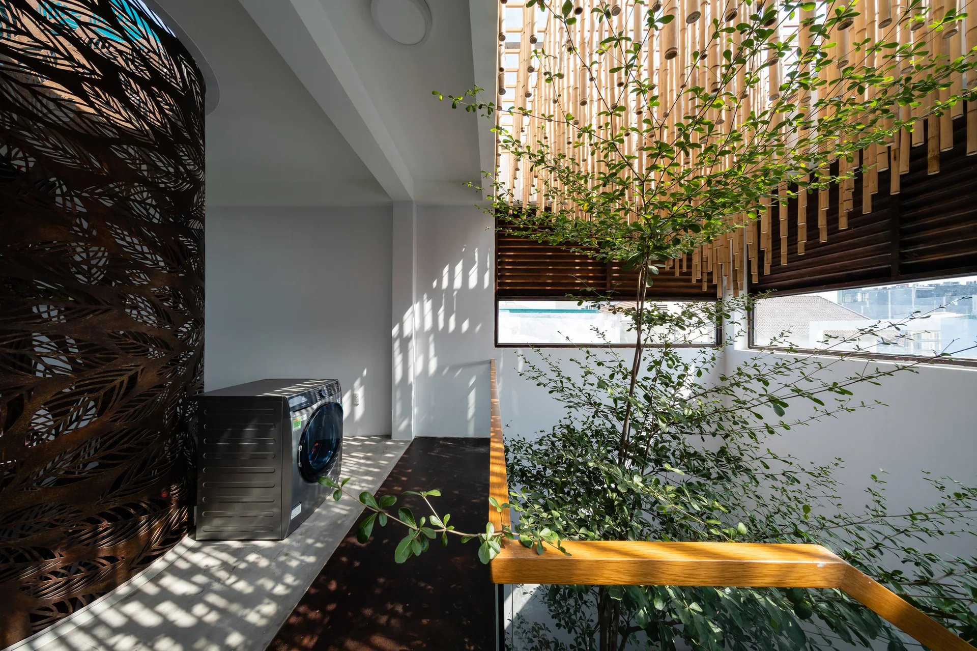A front-load washing machine sits on a concrete platform next to a tall indoor tree and a decorative laser-cut metal partition.