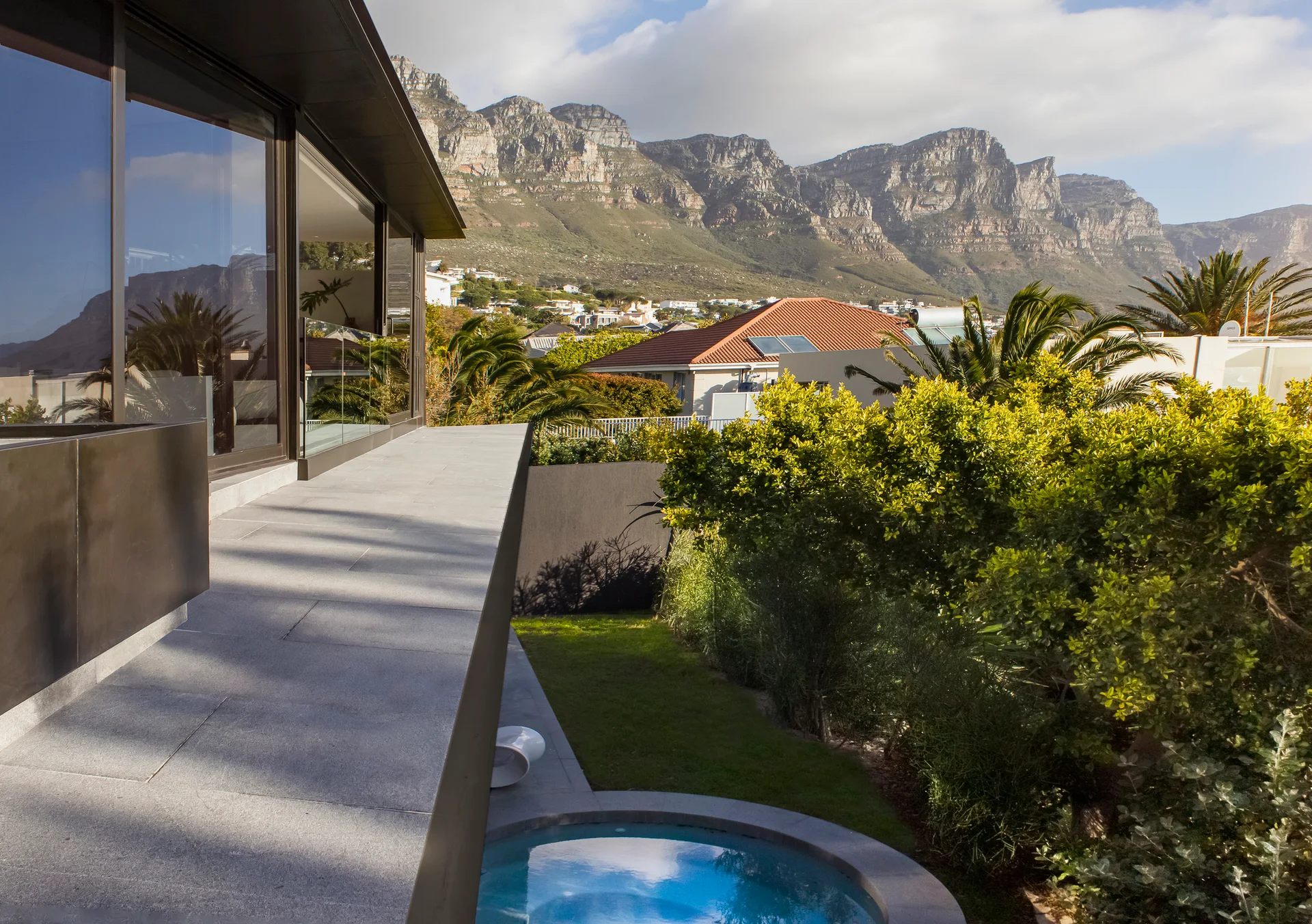 A modern grey stone terrace features a circular plunge pool set alongside lush green plants with mountains in the background.