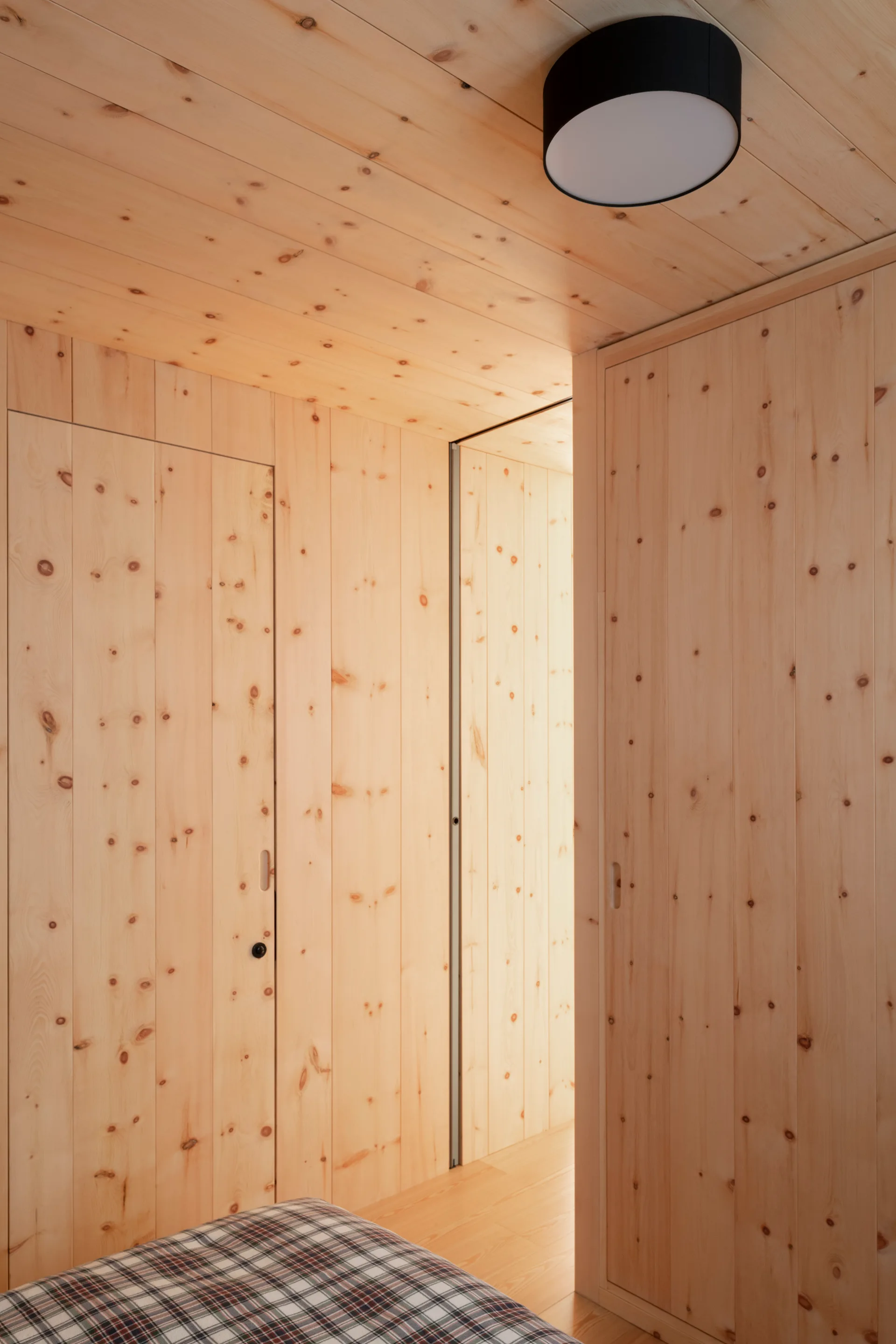 A minimalist bedroom interior with light knotty wood paneled walls, flush-integrated doors, and a round black ceiling light.
