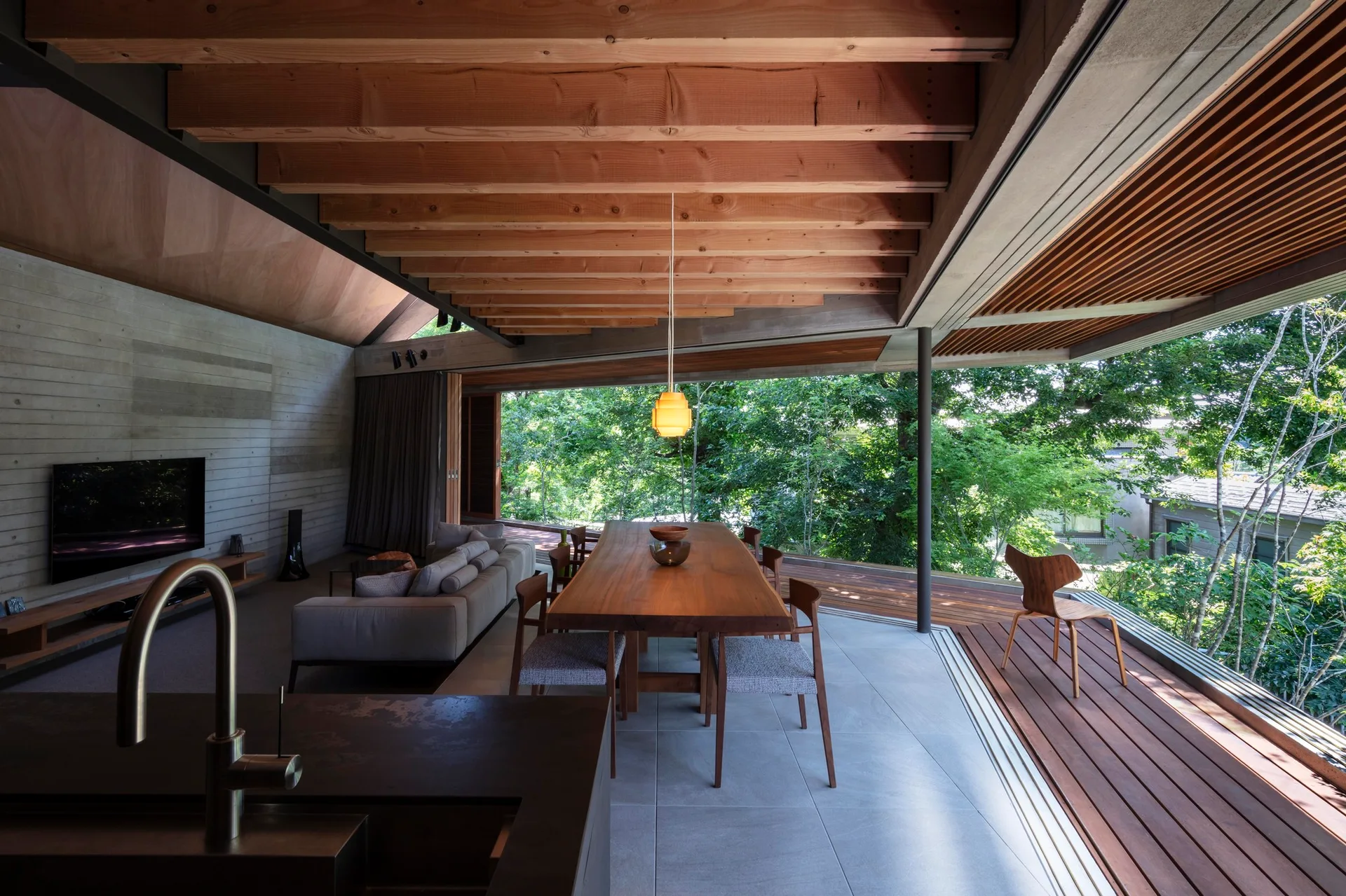 A modern open dining room features a long wooden table, concrete walls, exposed timber ceiling beams, and sliding glass doors that lead to an outdoor deck.