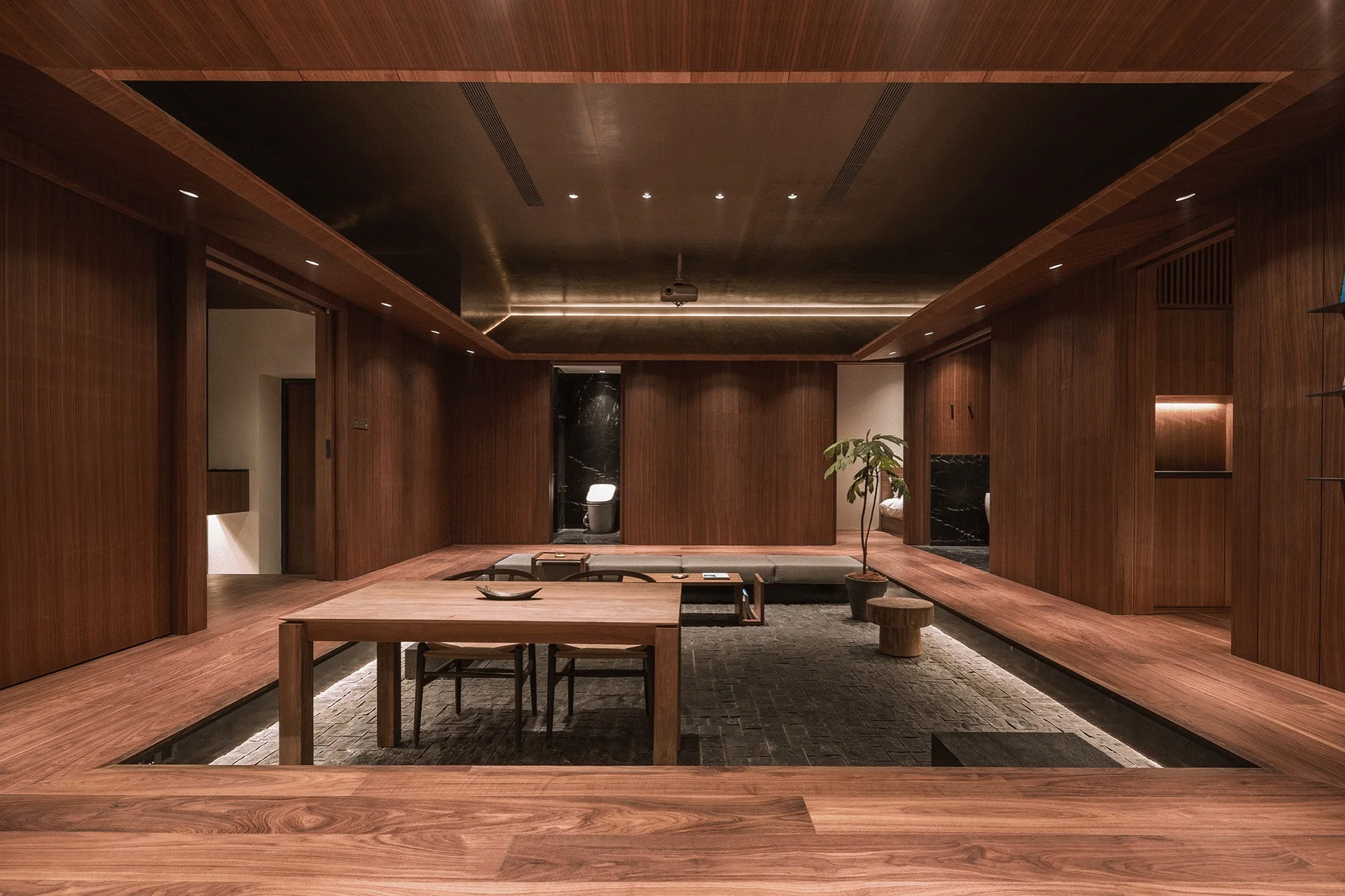 A minimalist sunken living room features a dark stone tile floor surrounded by warm wood-paneled walls and matching elevated platforms.
