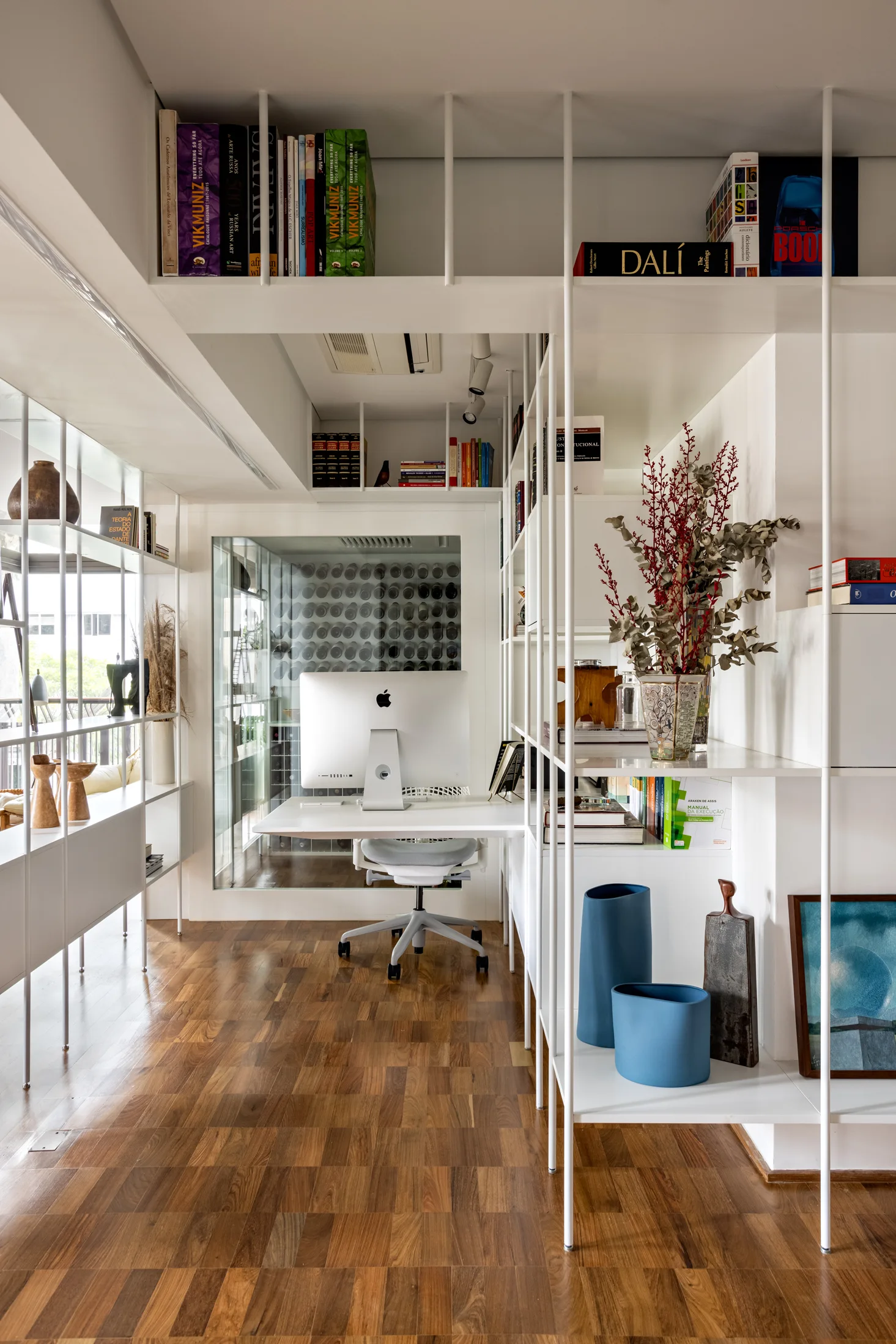 A minimalist workspace features a white desk, a grey office chair, and floor-to-ceiling white metal grid shelving on warm parquet flooring.