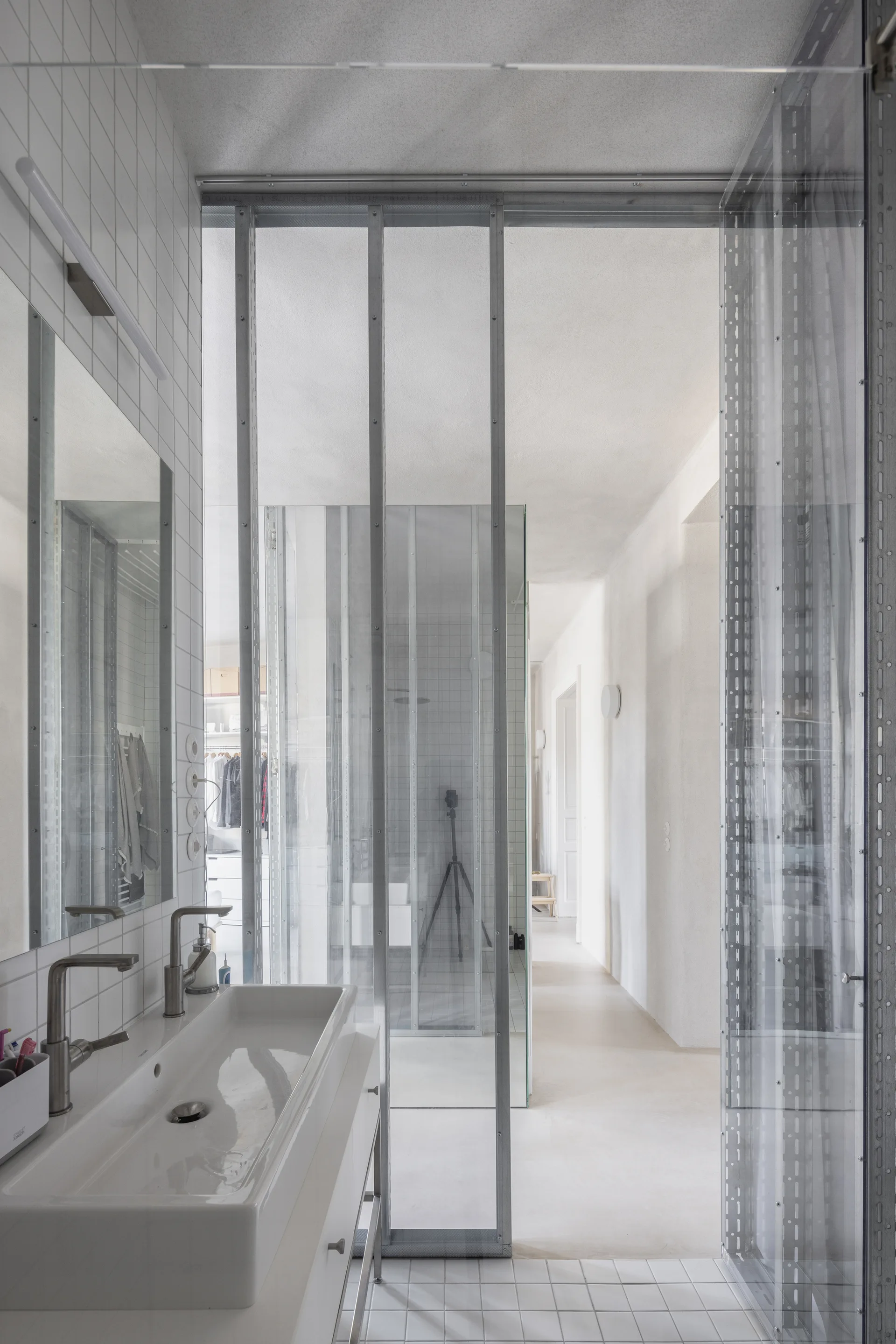 A minimalist white bathroom features a ceramic sink with a metal faucet, a vanity mirror, and an industrial glass partition framed in exposed metal studs.