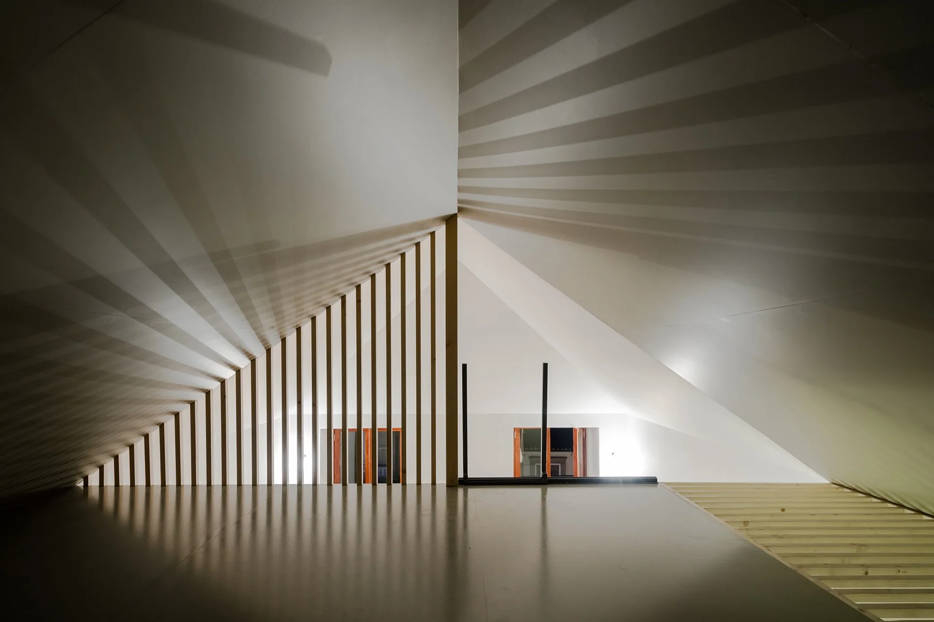 A minimalist loft landing features a sloped white ceiling and a vertical wooden slat balustrade casting linear shadows on the walls.