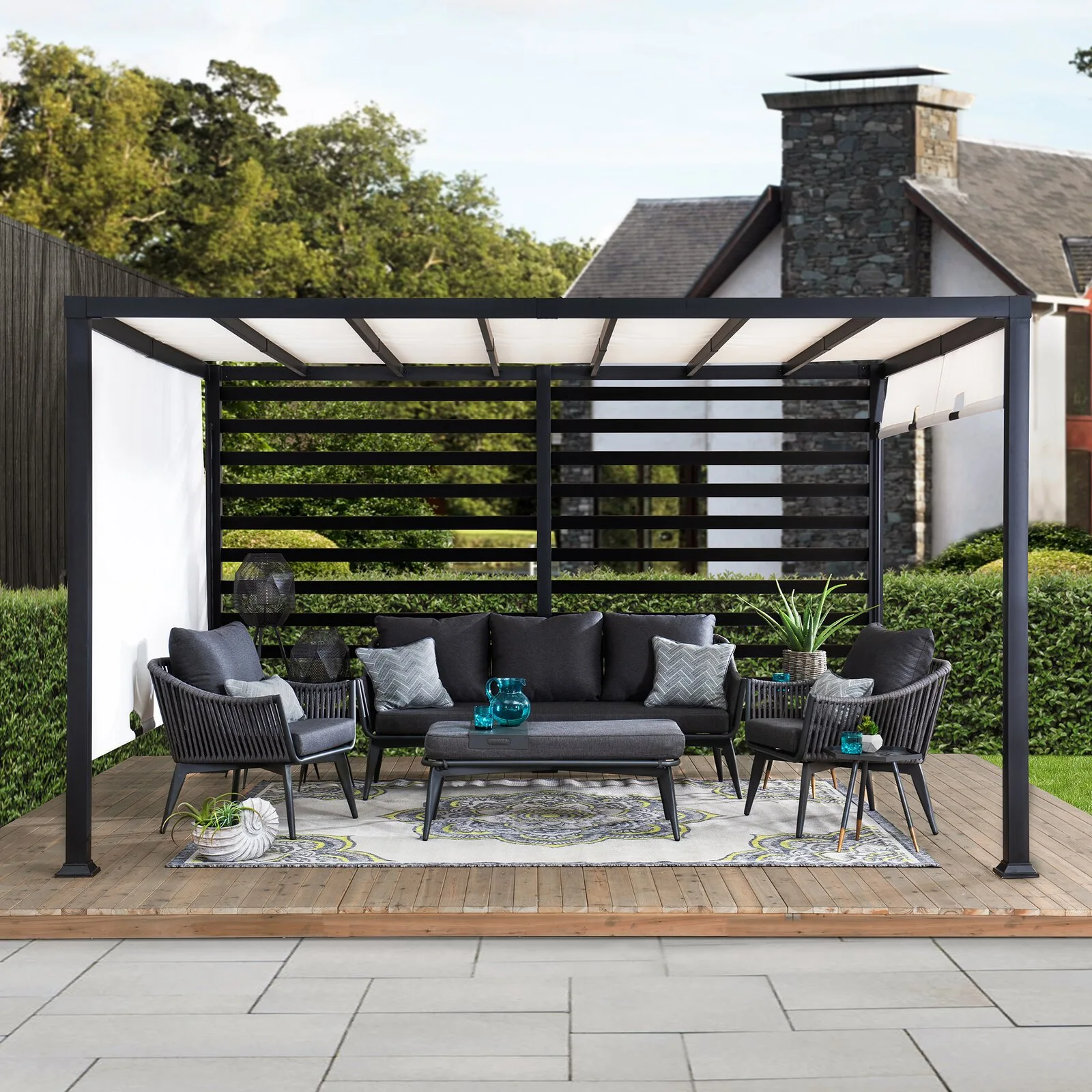 A black metal pergola houses a dark gray outdoor sofa set on a wooden deck with a patterned area rug and a slatted privacy screen.