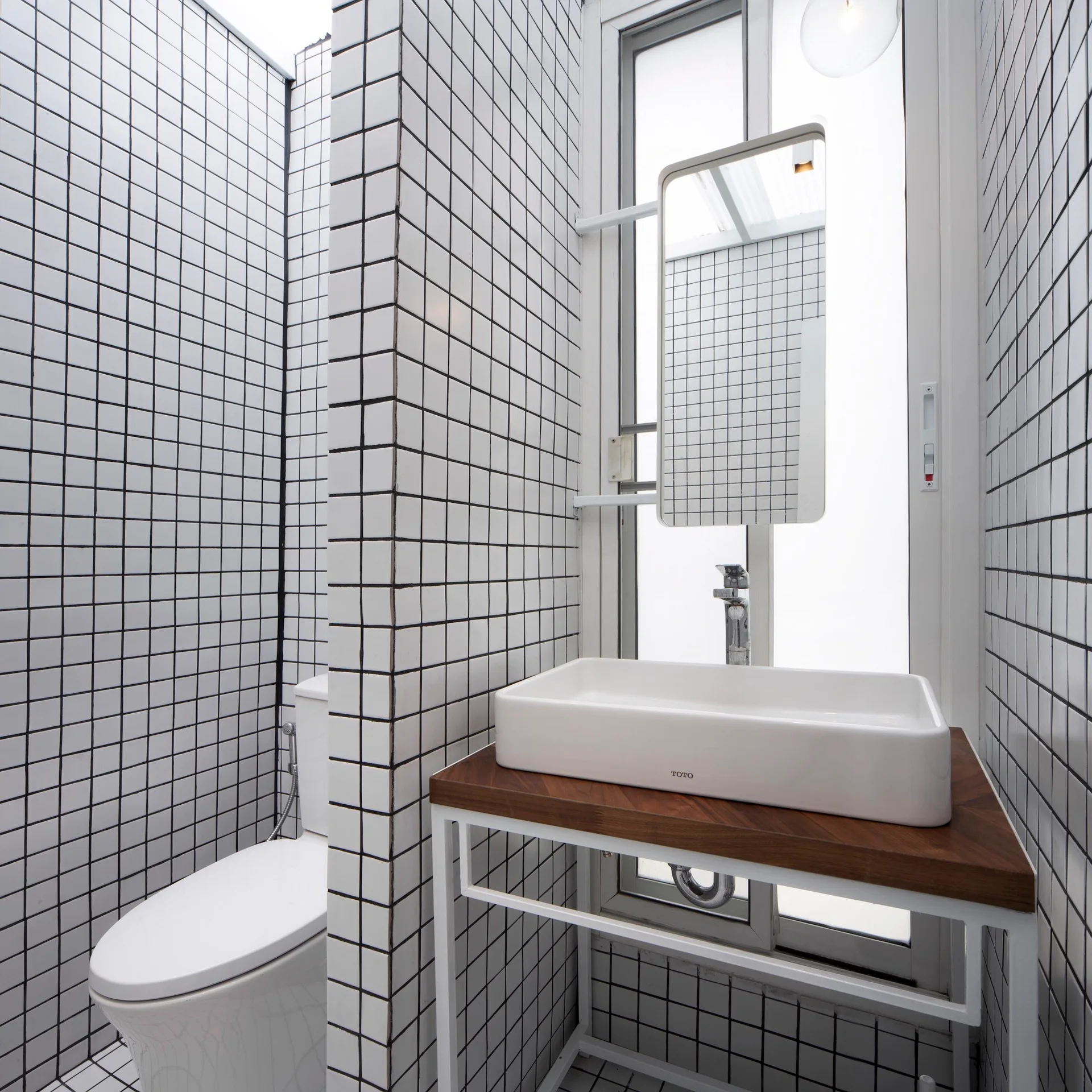 A modern bathroom features floor-to-ceiling white grid tiles with black grout, a white toilet, and a wood-topped metal vanity with a vessel sink.
