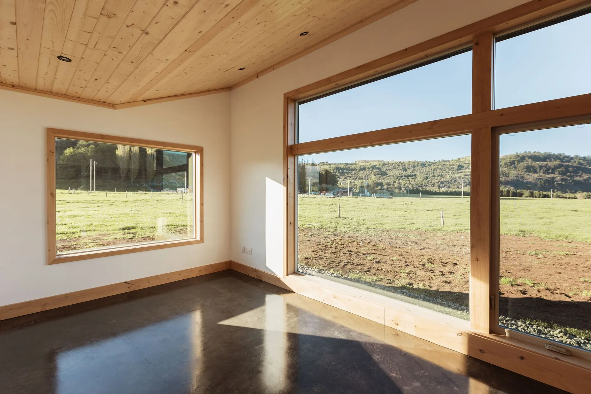 An empty minimalist room features a sloped wood plank ceiling, white walls, large wood-framed windows, and a polished dark concrete floor.