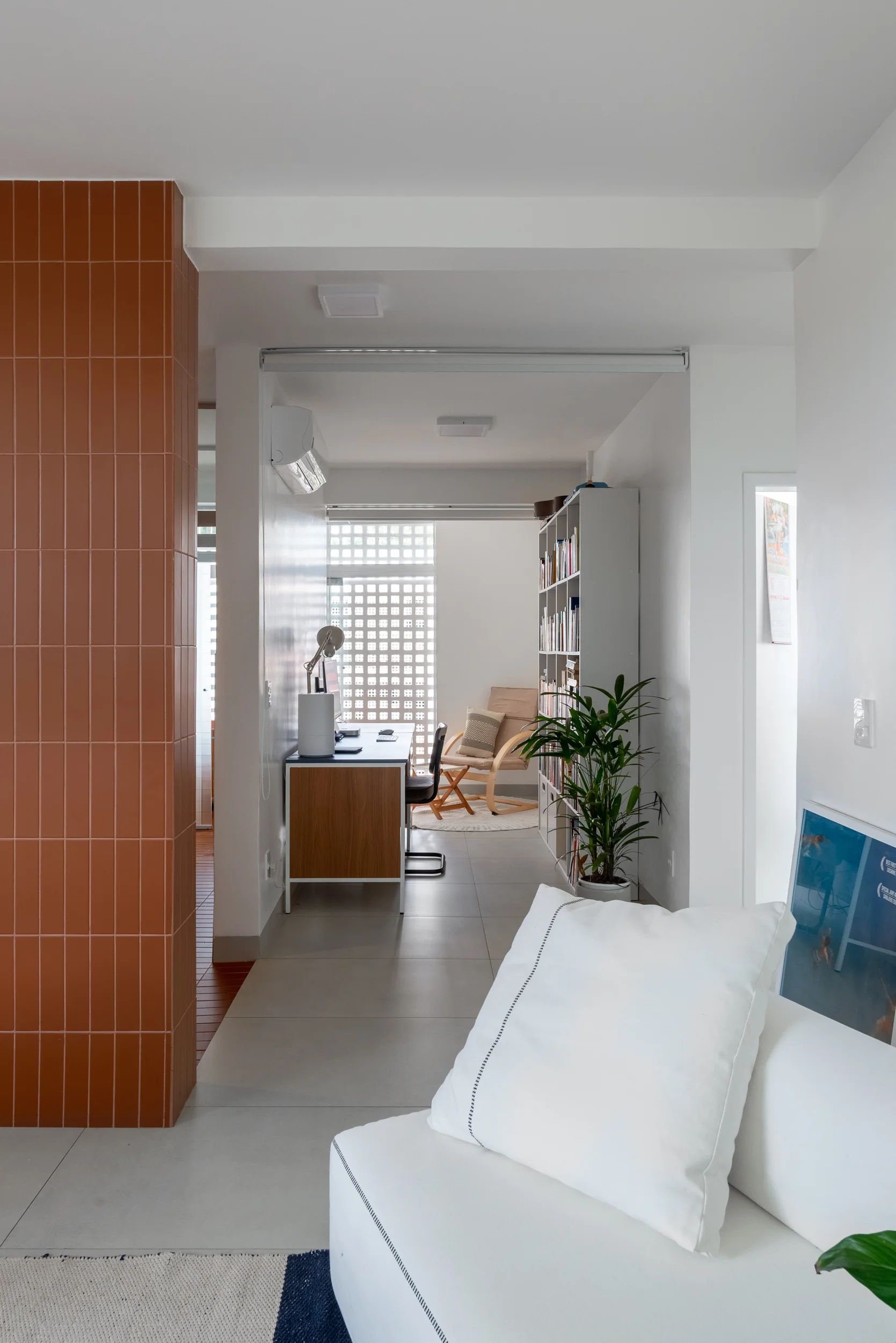 A bright living room features a terracotta tiled column in the foreground leading to a home office with a desk and a breeze block wall.
