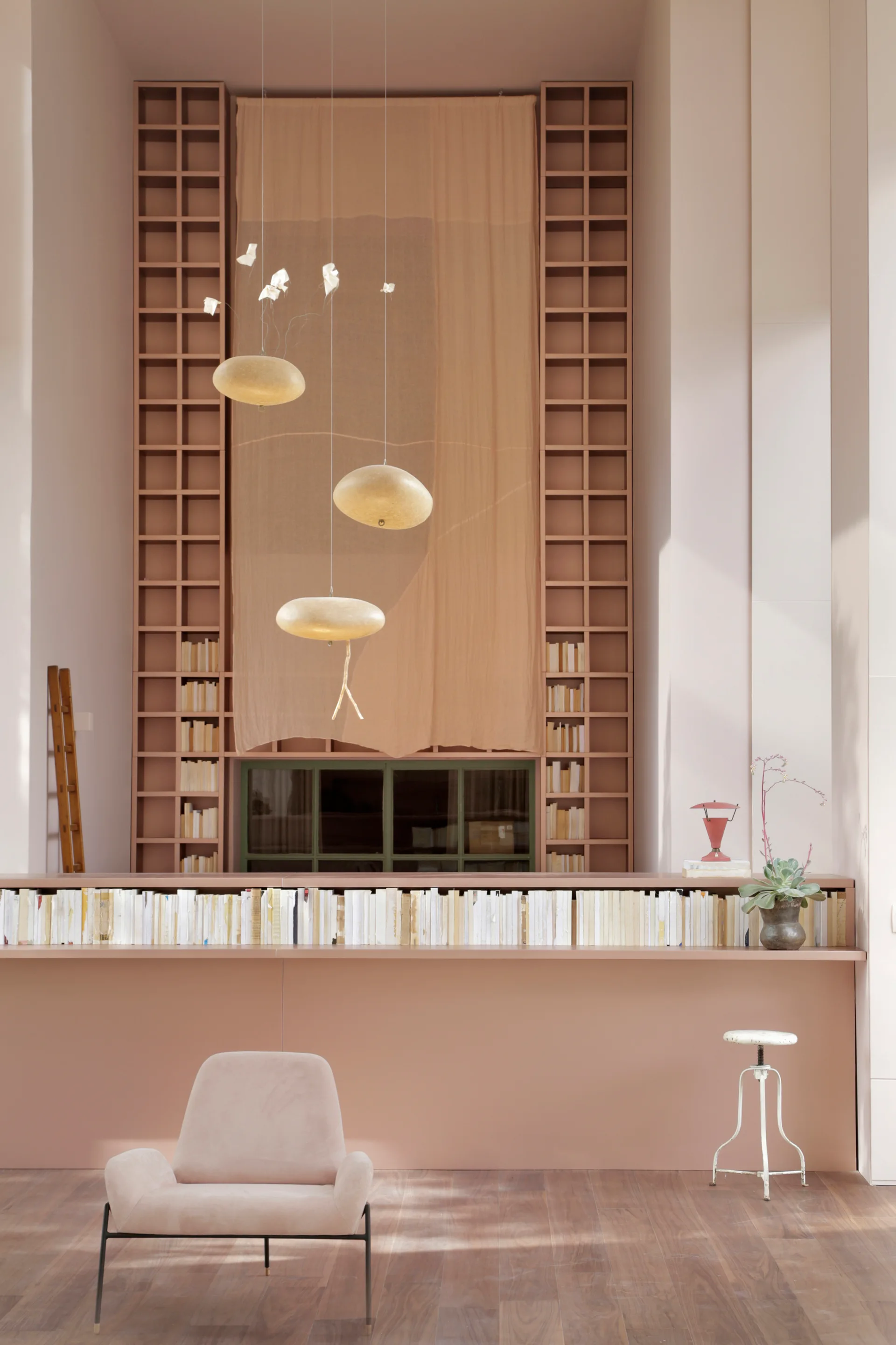 A tall dusty pink living room features symmetrical built-in bookcases, a row of books with page-sides facing out, and three gold pendant lights.